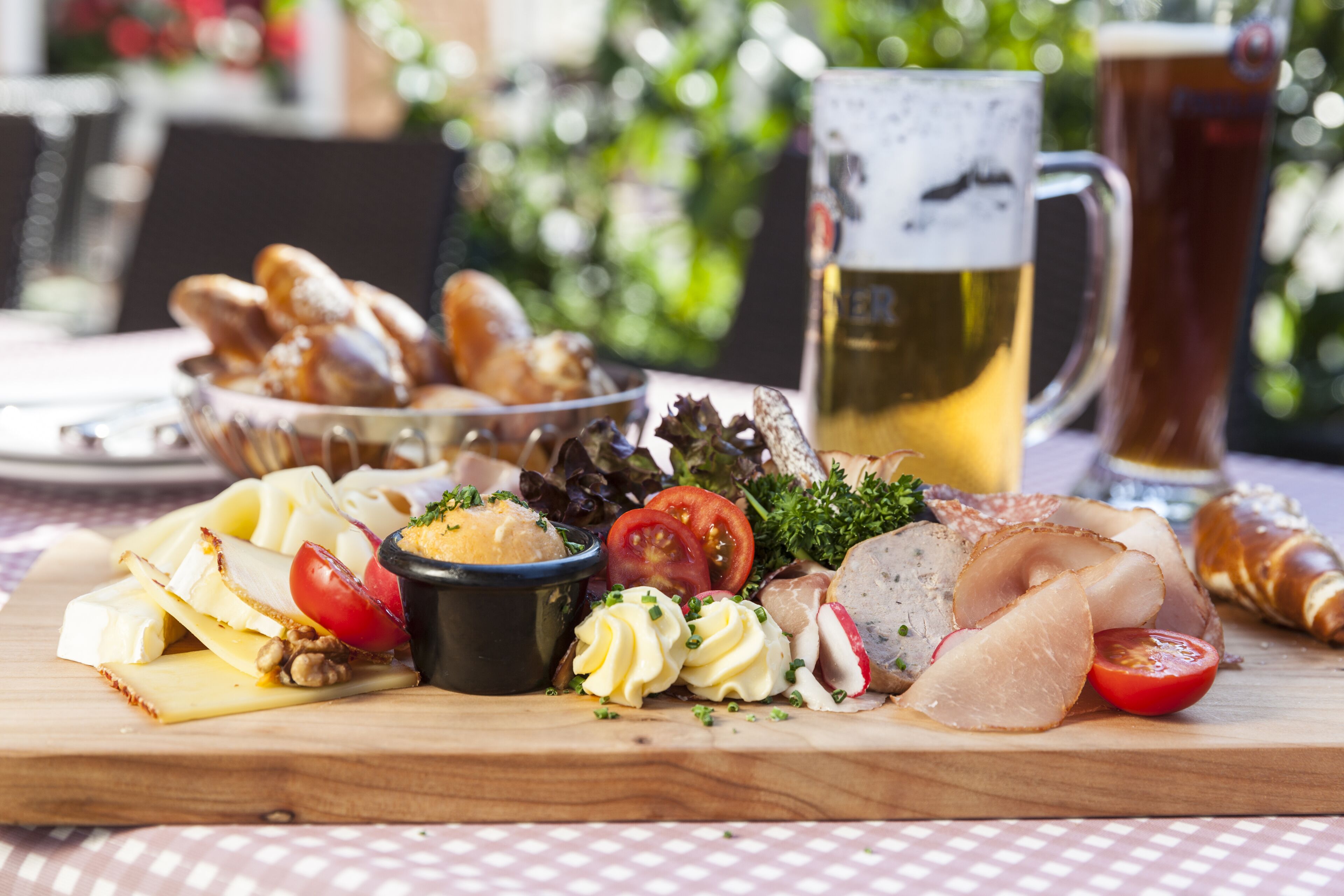 A charcuterie board with various meats, cheeses, and vegetables on a wooden board, accompanied by a mug of beer and a glass of beer in the background.