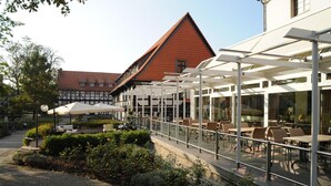 An outdoor cafe area with tables and chairs, a covered walkway, and a building with a red roof in the background. - Hotel Ratskeller (Salzgitter)