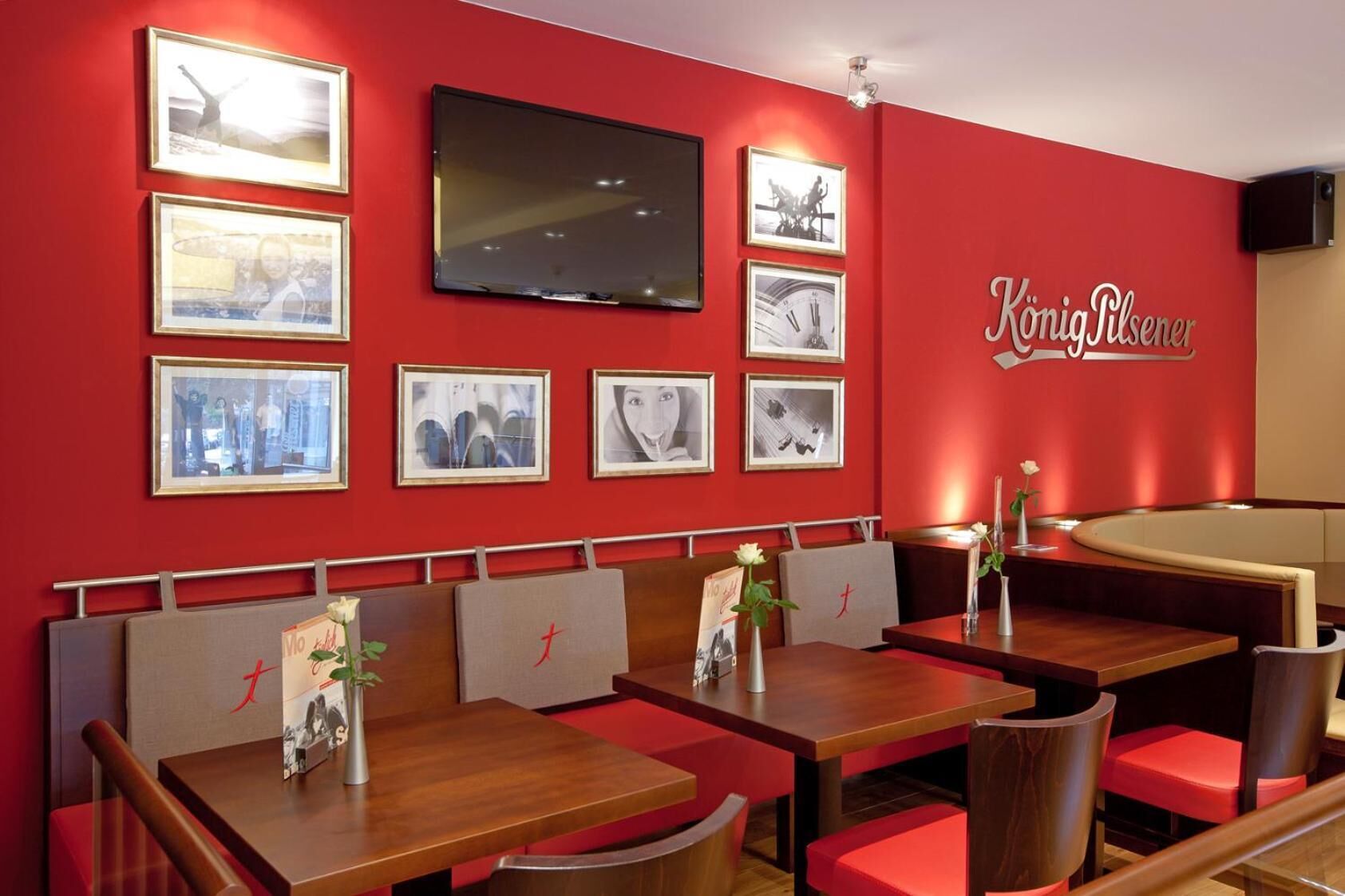 A restaurant interior with red walls, multiple framed pictures, a TV mounted above the seating area, and a sign reading "Königa Pilsener".