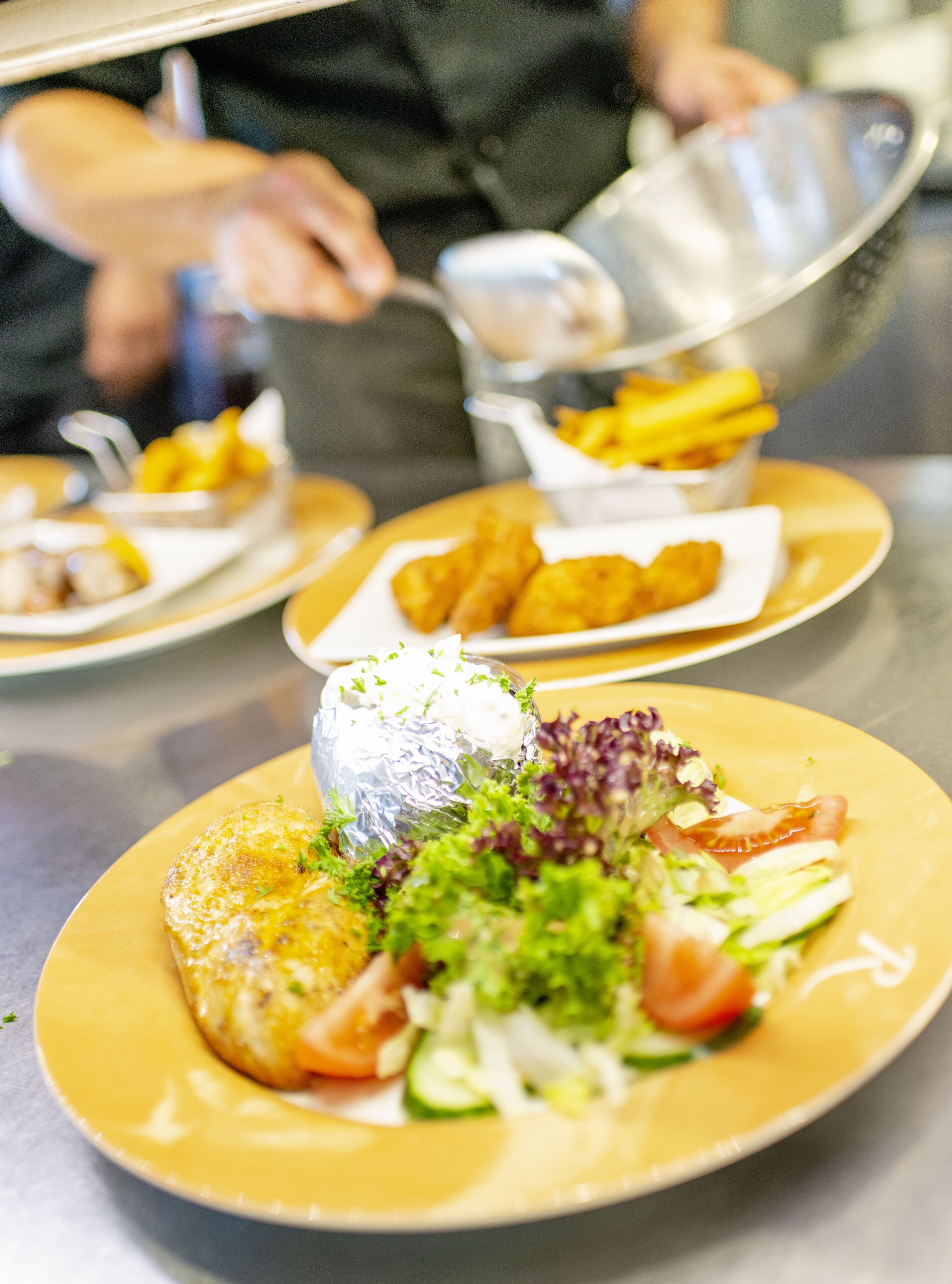 A dining table with various dishes, including a salad with greens and a fish fillet, and a person serving food.