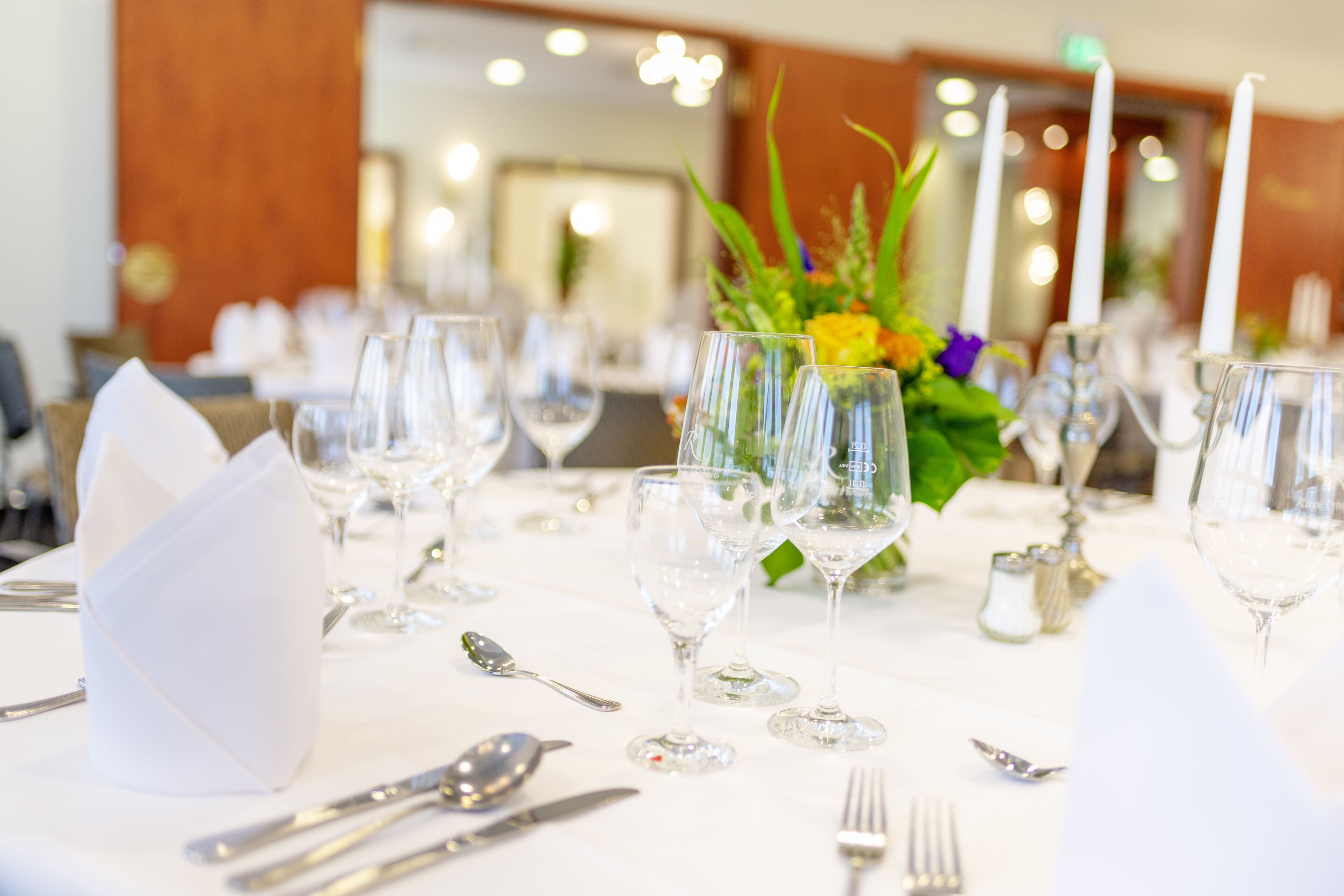 Una mesa lista para comer, con cristalería transparente, cubiertos y un centro de mesa con flores.