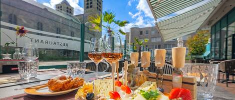 Un área de comedor al aire libre con una variedad de quesos, frutas y copas de vino, con vista a un edificio en el fondo.