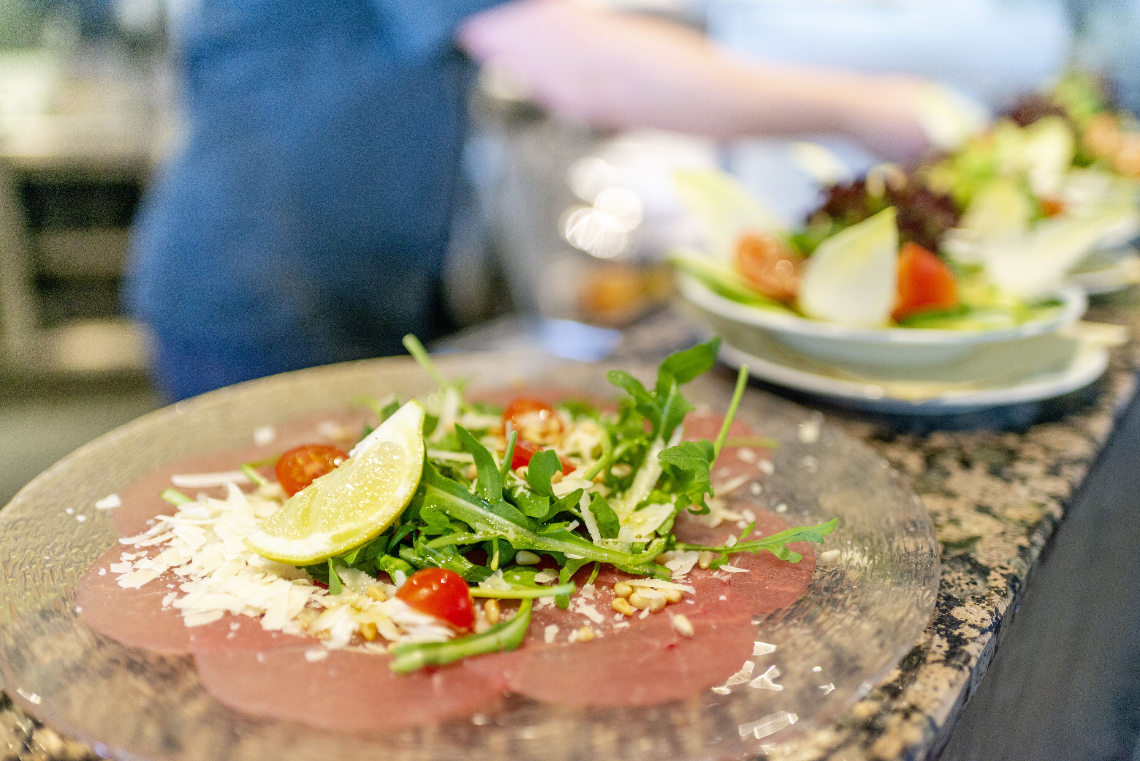 A close-up of a dish with thinly sliced meat, greens, and a slice of lime on a glass plate.