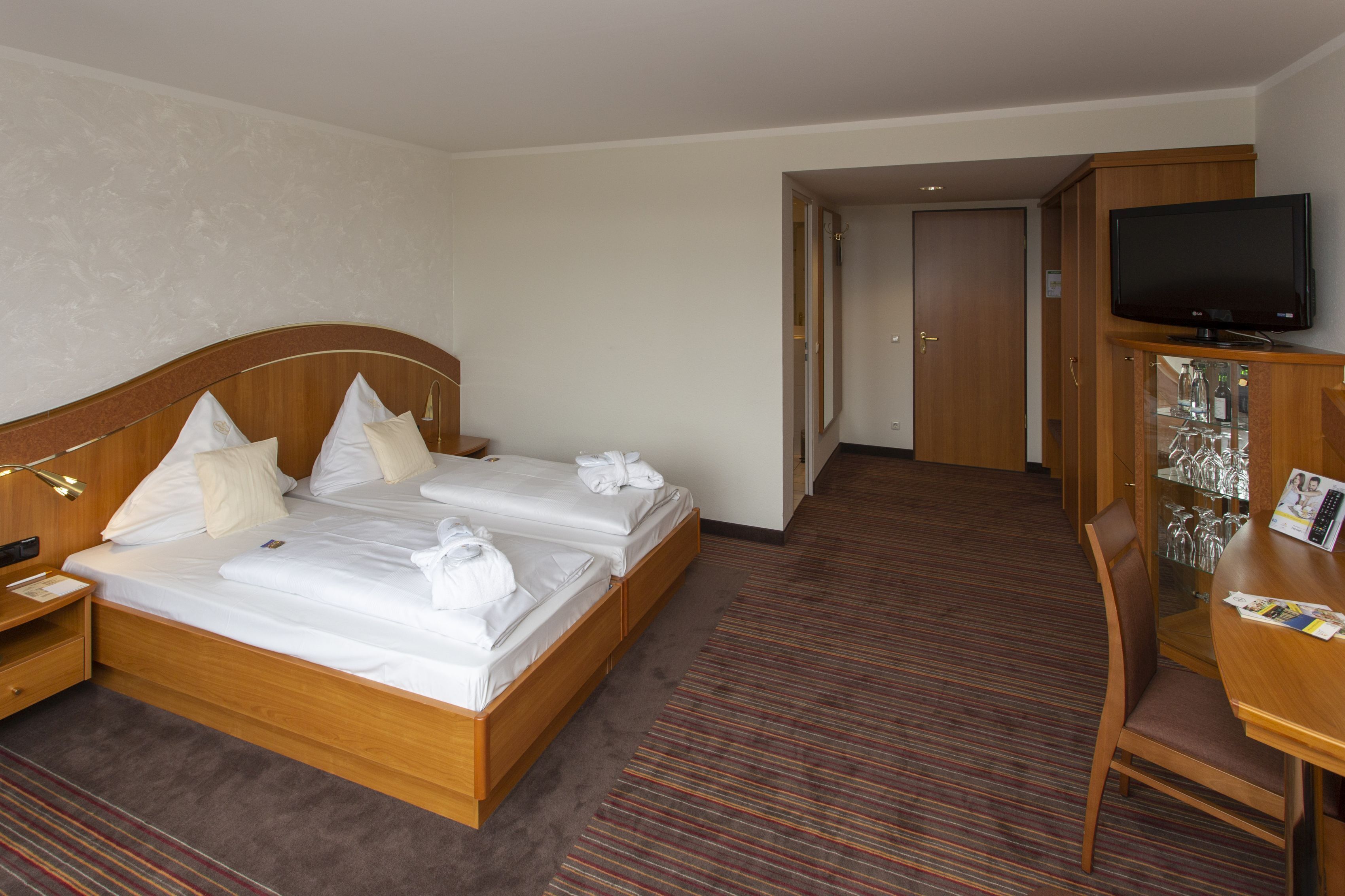 A hotel room with a double bed, bedside tables, a TV, a desk with a chair, and a glass cabinet with bottles.