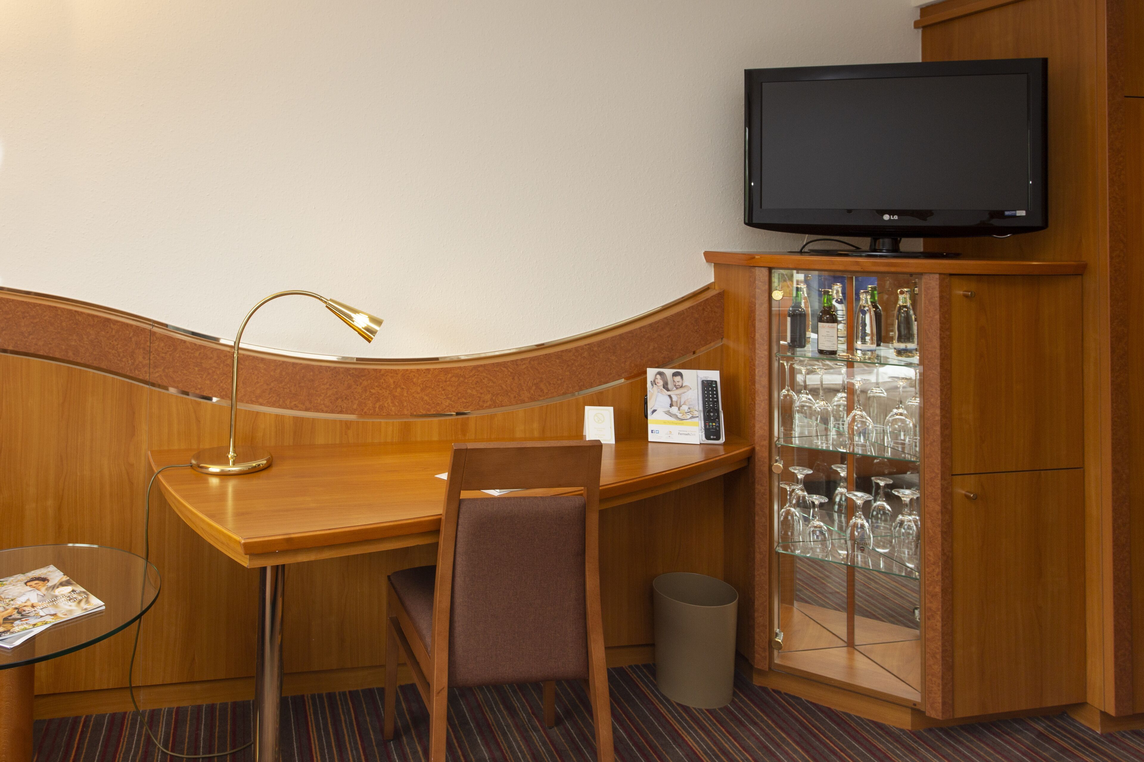 Quarto de hotel com uma escrivaninha de madeira, televisão, cadeira, mesa de vidro e um armário com copos.