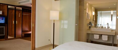 Una habitación de hotel con una cama grande, cabina de ducha de vidrio, tocador con espejo y un televisor en la pared izquierda.