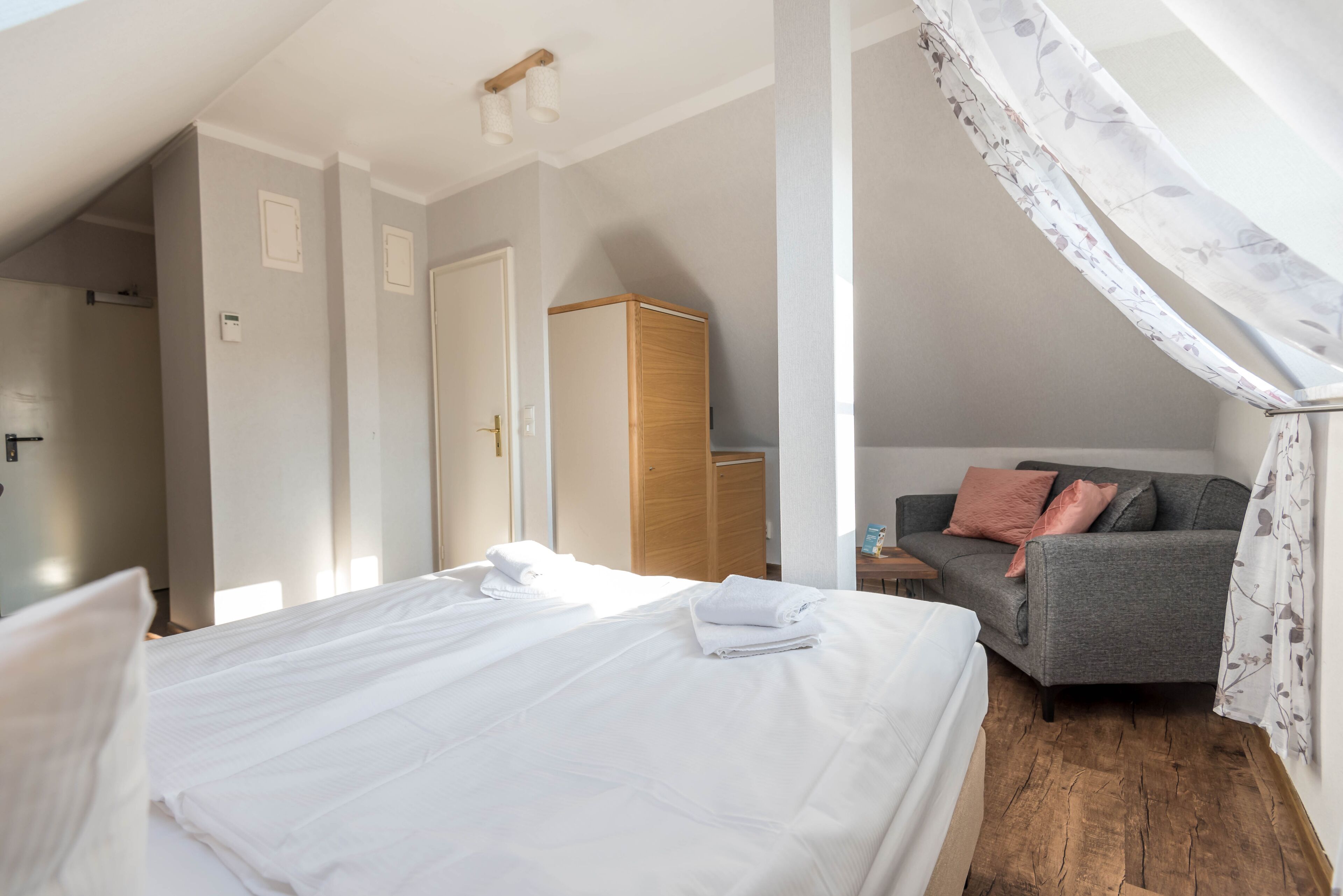 A bedroom with a neatly made bed, a grey sofa with cushions, a wooden wardrobe, and a window with a floral curtain.