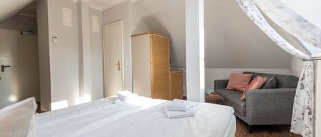 A bedroom with a neatly made bed, a grey sofa with cushions, a wooden wardrobe, and a window with a floral curtain.