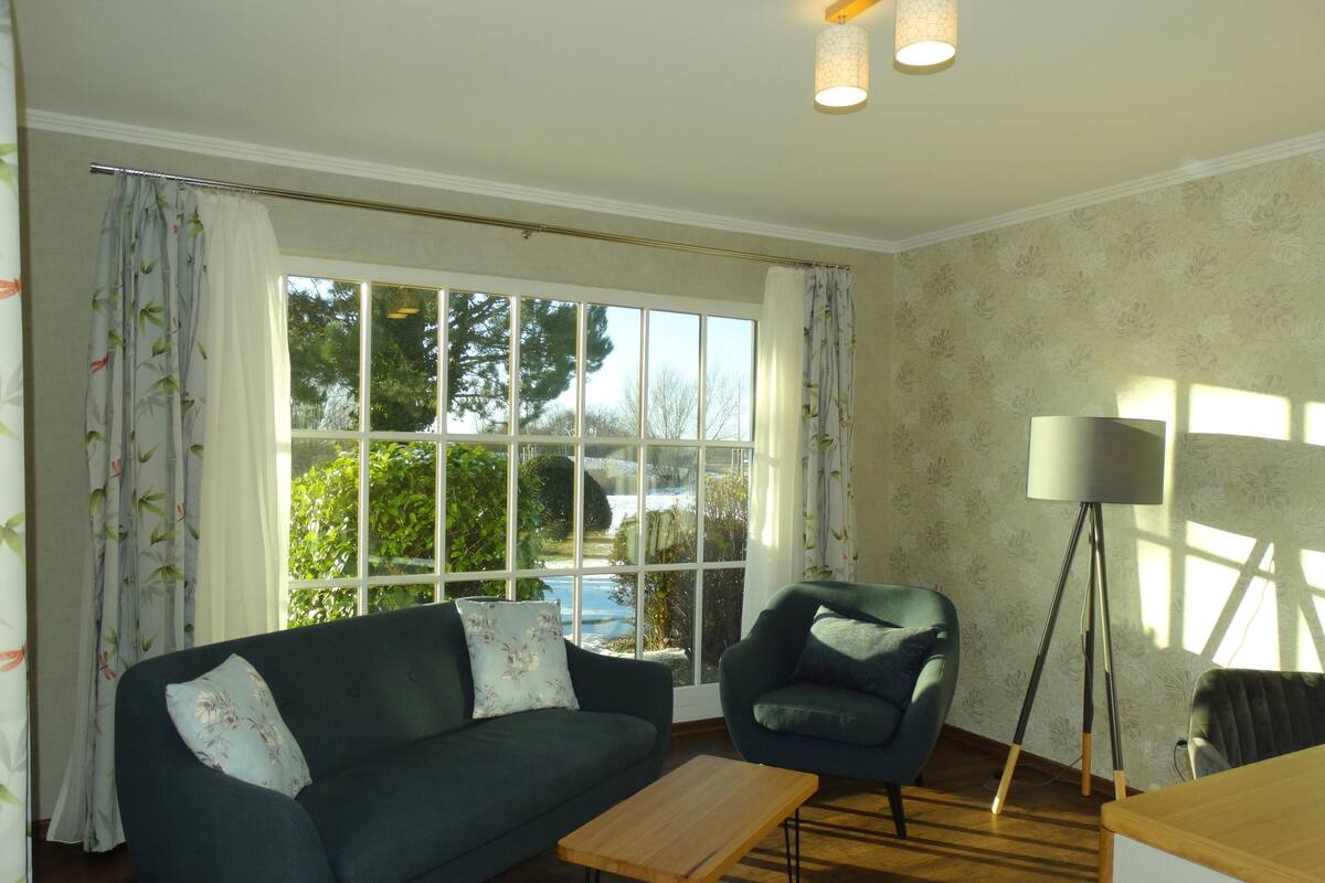 A living room with a sofa, armchair, and a small table. There is a window with curtains, a floor lamp, and a view of greenery outside.