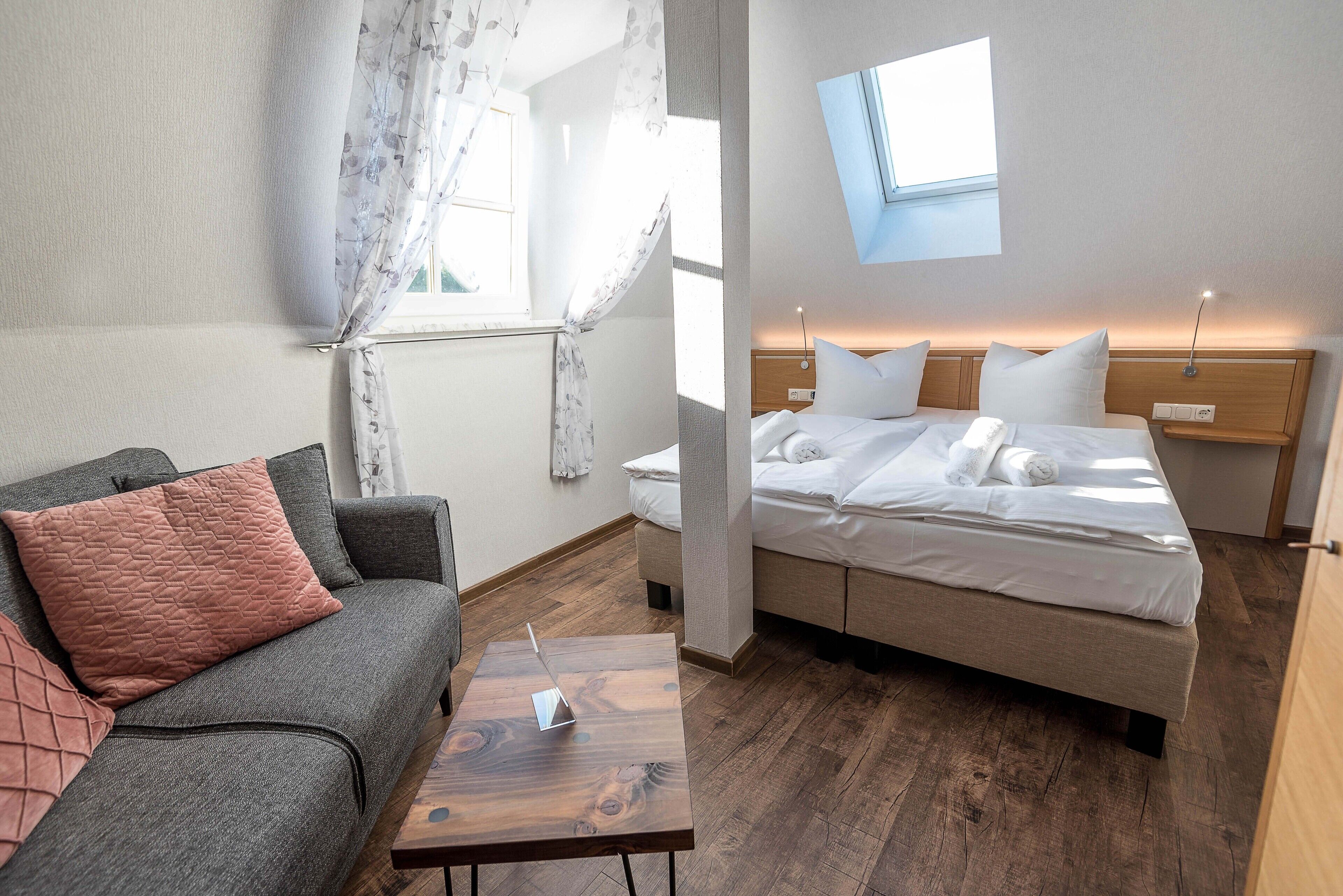 A modern hotel room with a grey sofa, a wooden coffee table, a bed with white bedding, and a skylight above the bed.