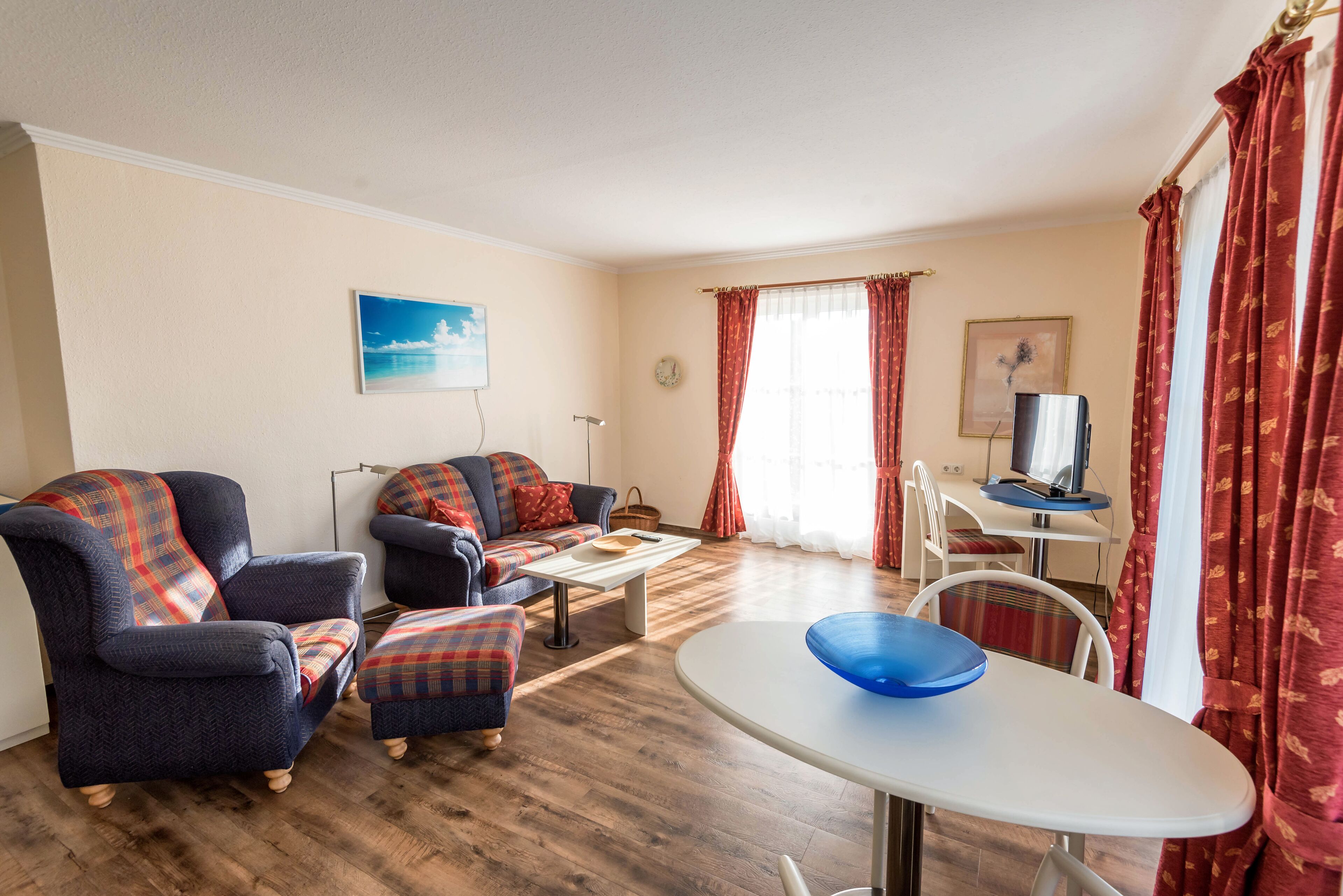 Een woonkamer met een bank, fauteuil, pouf, een eettafel met een blauwe schaal, en een televisie op een bureau.