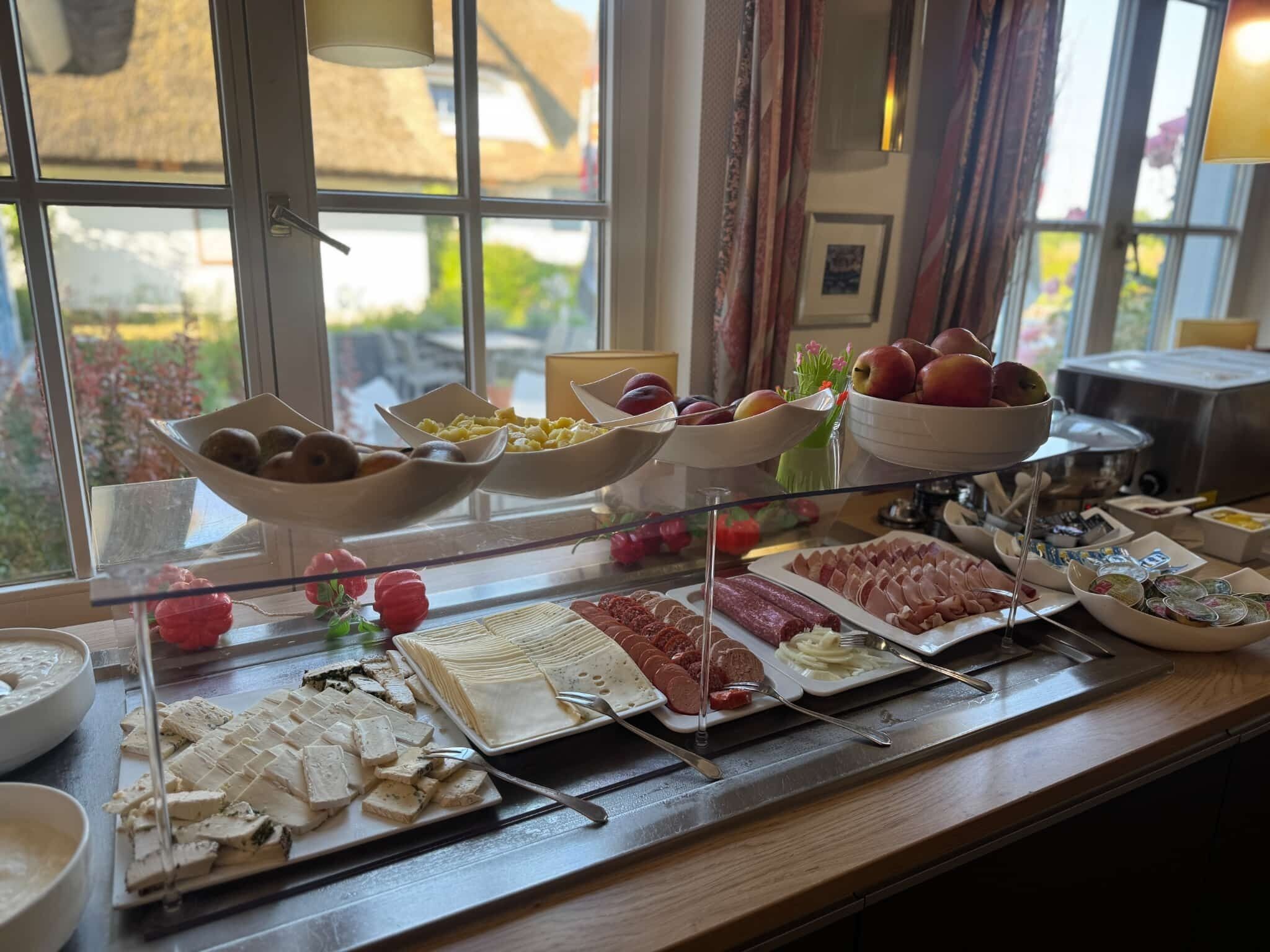En buffé med ett varierat utbud av mat, inklusive frukt, ostar och charkuterier, som presenteras på en glasskiva nära ett fönster med utsikt över grönområden.
