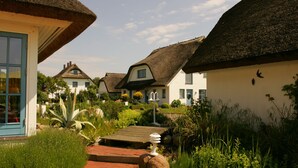 A row of traditional houses with thatched roofs, a garden with various plants, and a wooden pathway leading to the entrance.