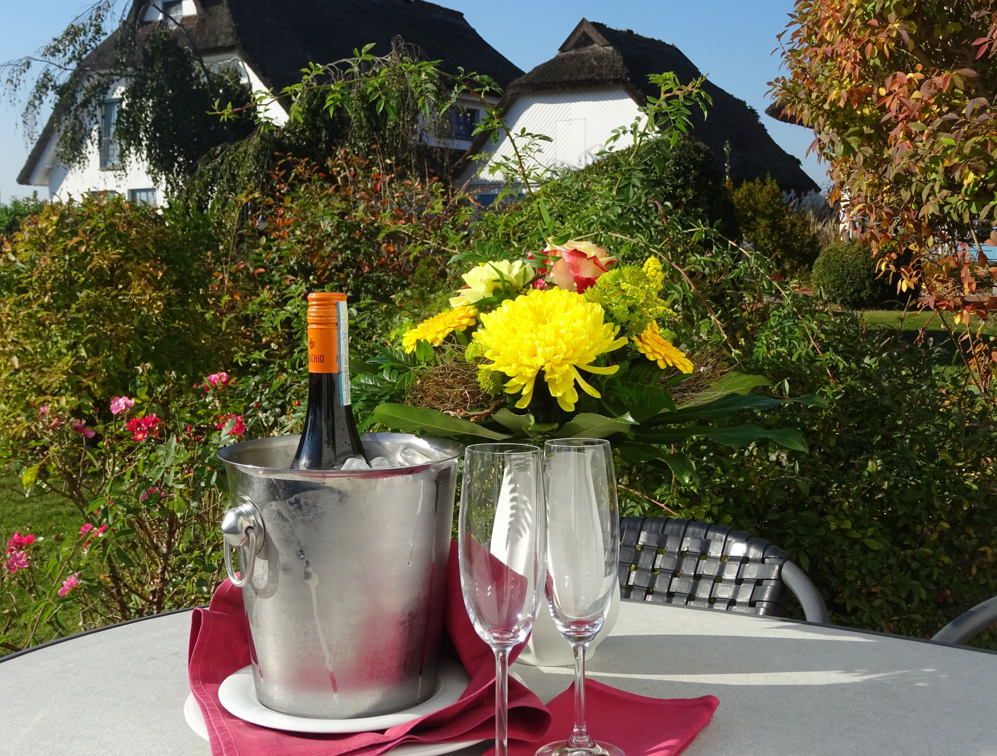 A bottle of wine in an ice bucket on a table with two glasses, surrounded by flowers and greenery.