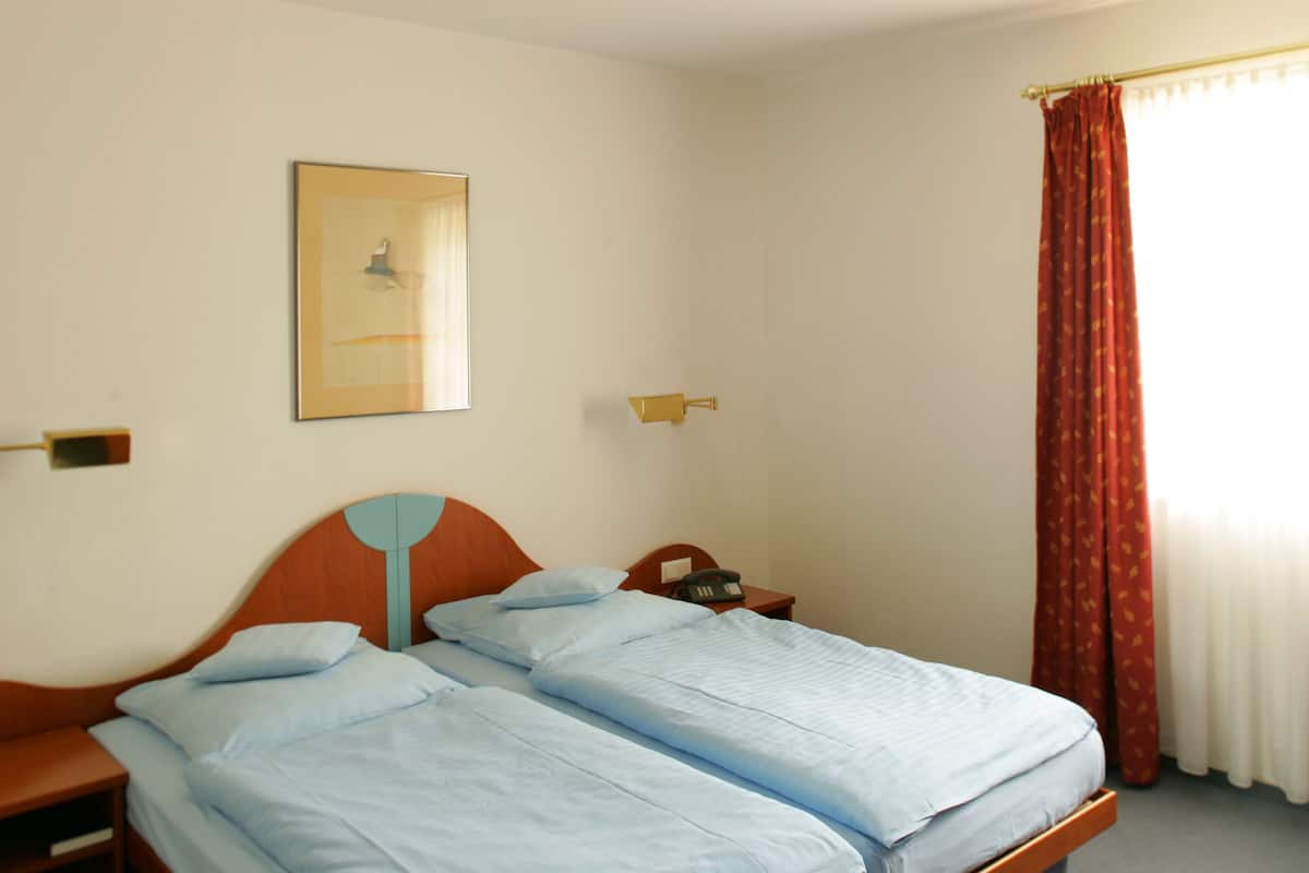 A hotel room with a double bed, a bedside table, a telephone, a wall-mounted lamp, and a window with curtains.