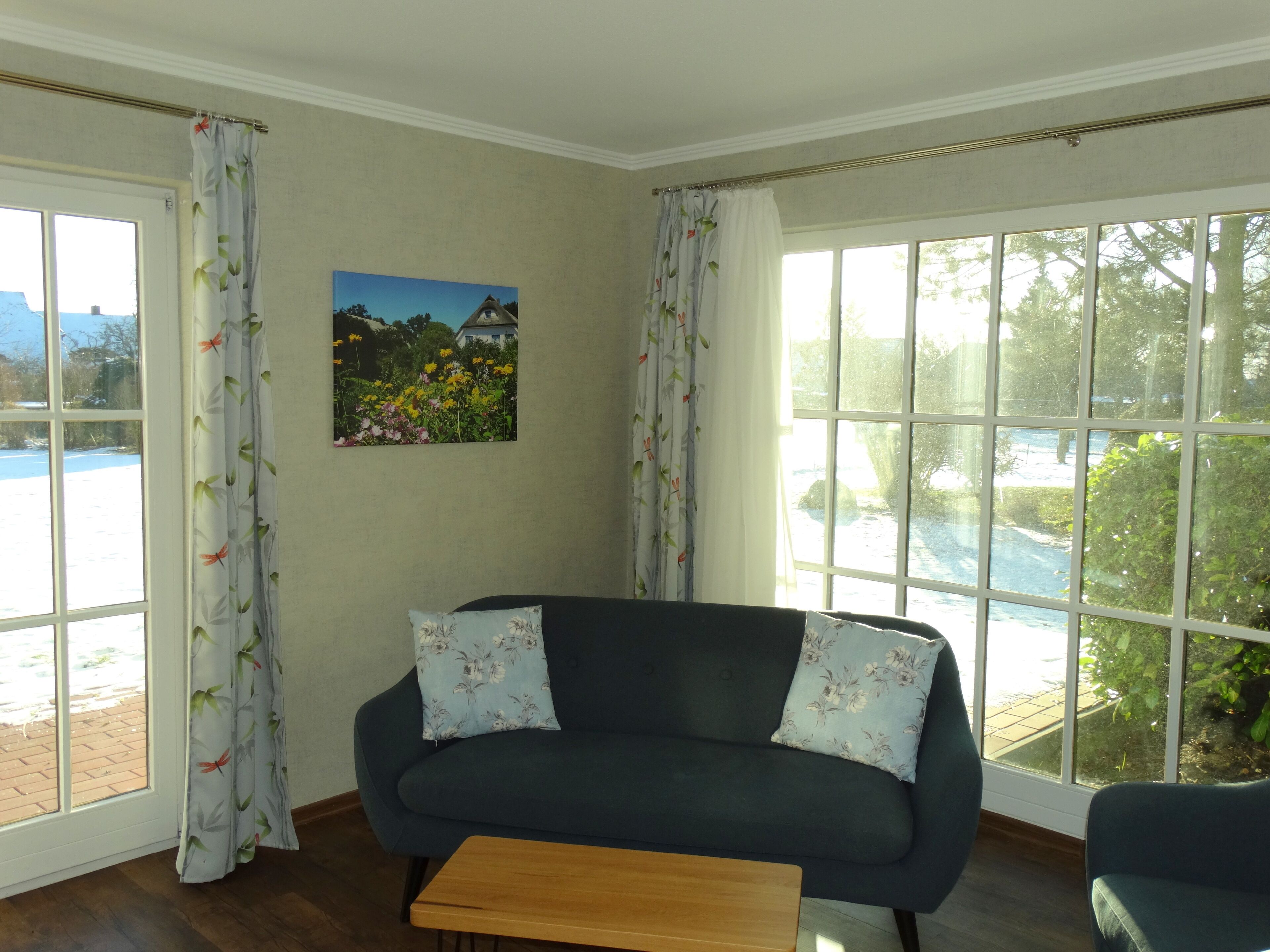 A room with a dark blue sofa, two floral cushions, a wooden coffee table, and a large window with a view of a body of water and greenery.