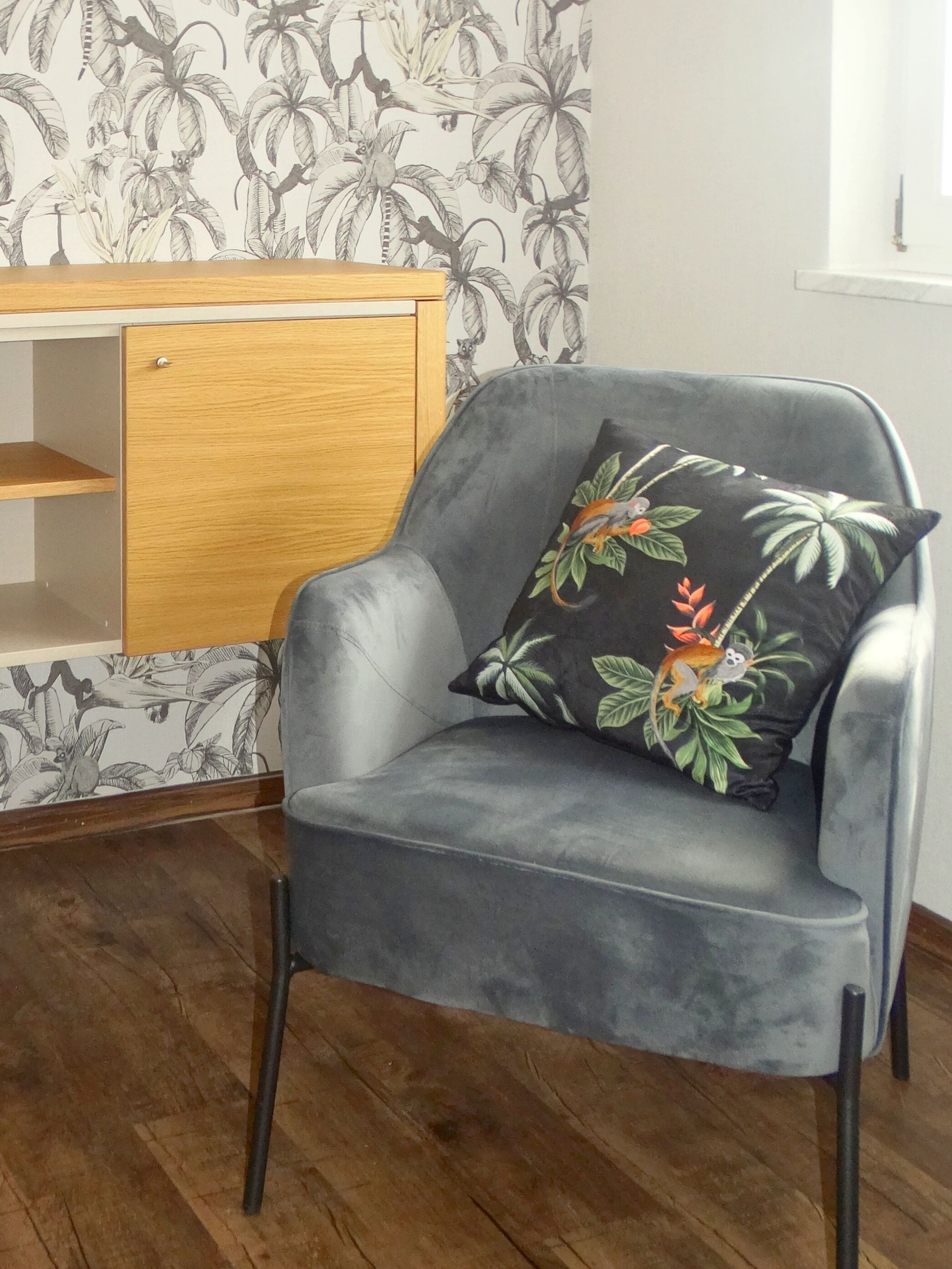 Ein grauer Sessel mit einem floralen Kissen, eine Holzkommode und eine Wand mit tropischem Muster.