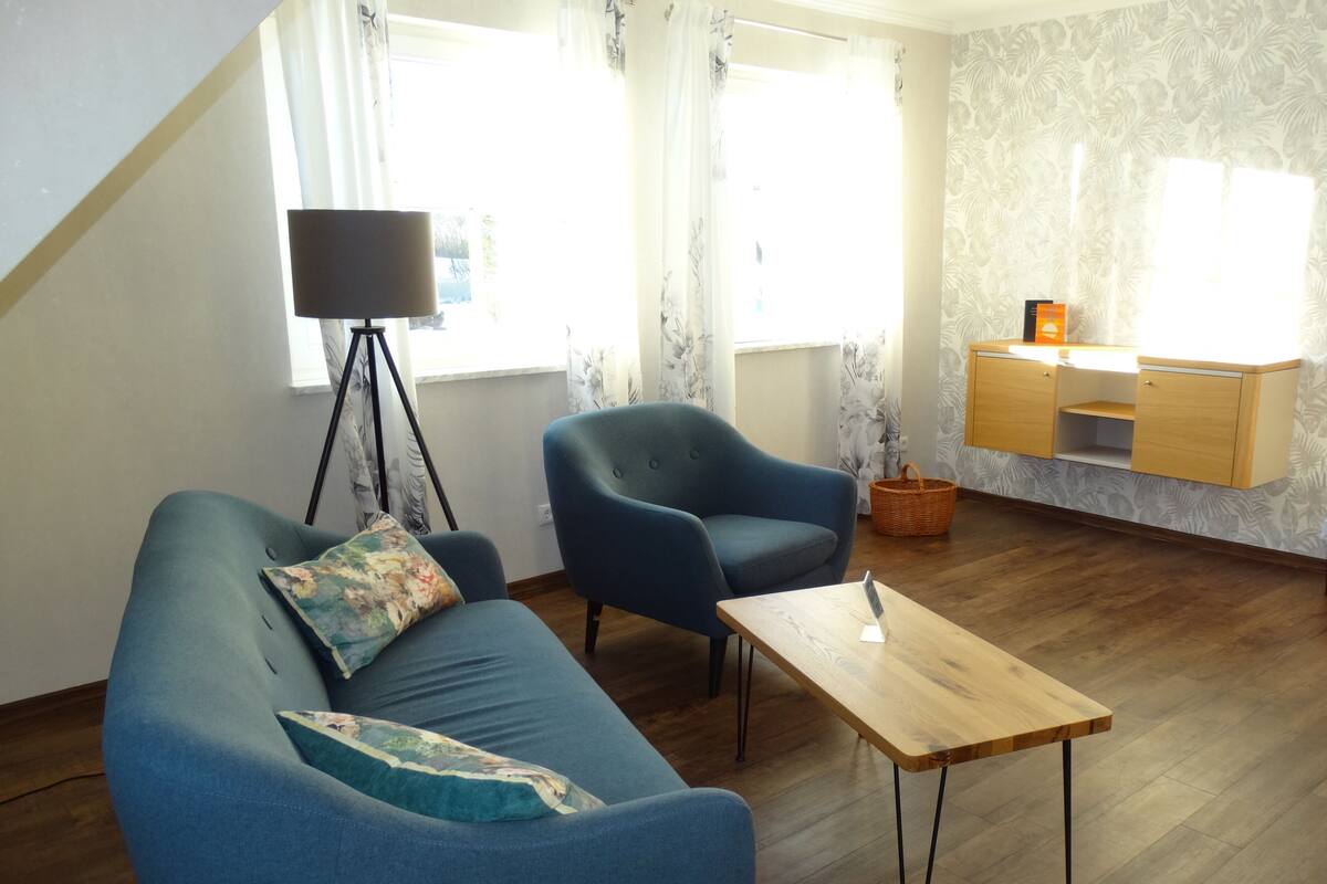 A living room with a blue sofa, a matching armchair, a wooden coffee table, a floor lamp, and a small desk with a basket.
