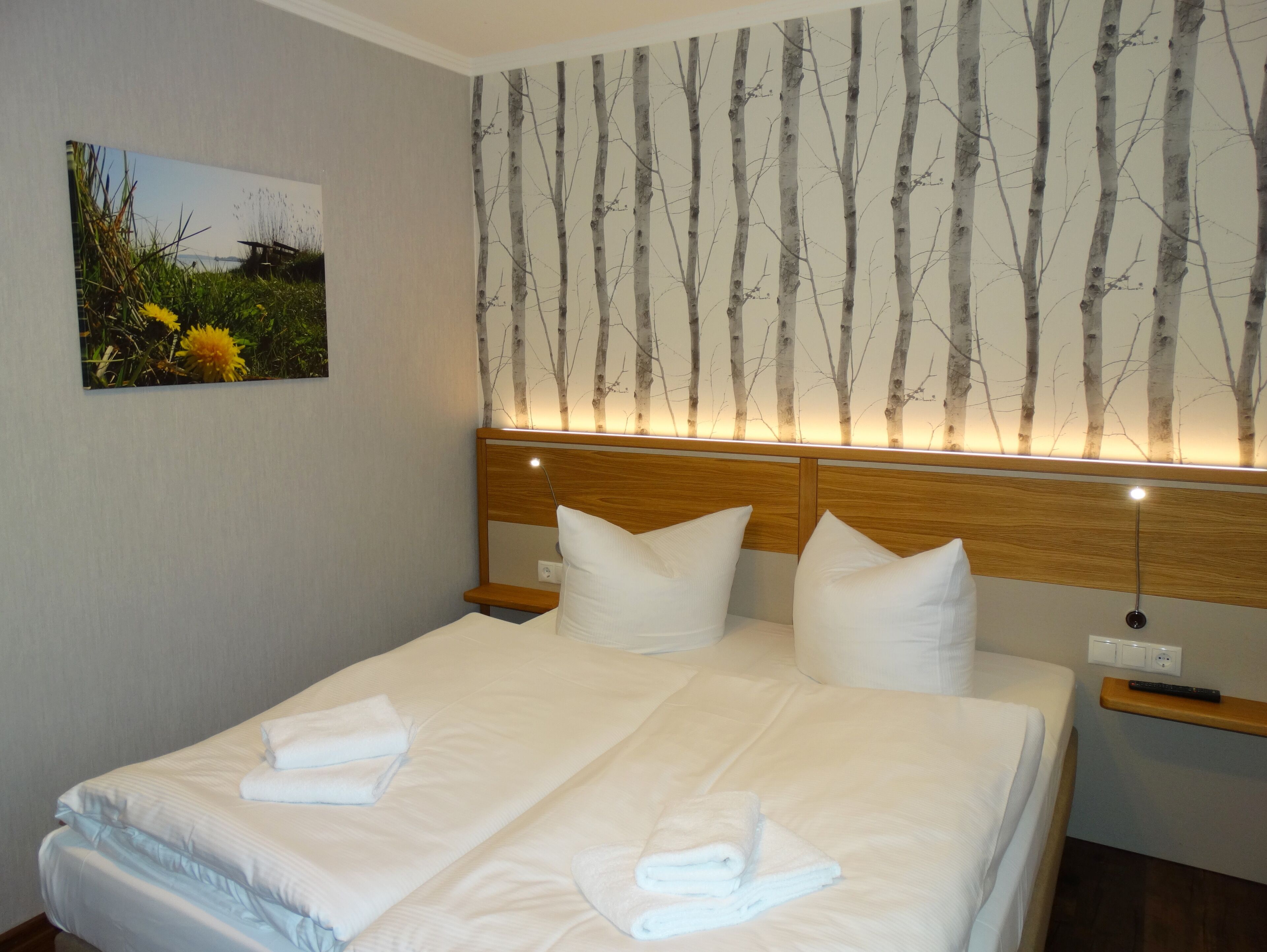 A double bed with white linens and two pillows, a bedside table with a lamp, and a wall with a nature-themed wallpaper.