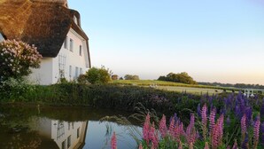 A white building with a thatched roof beside a calm river, surrounded by vibrant flowers and greenery.