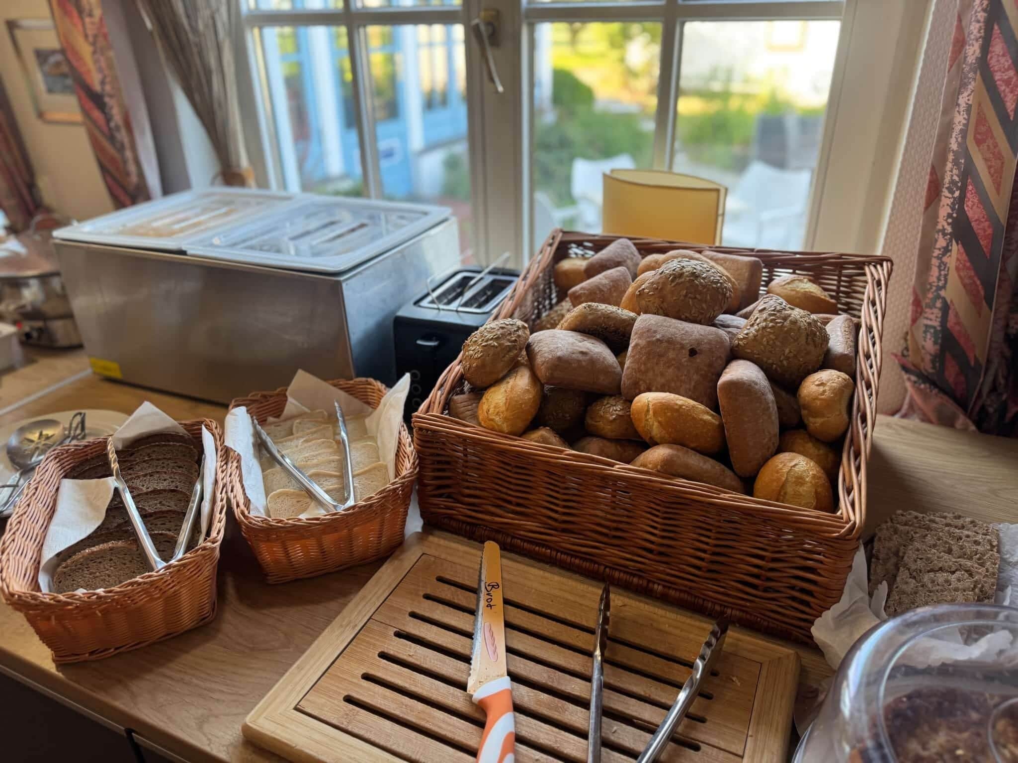 A breakfast buffet with various breads and pastries, a toaster, and a bread knife on a wooden counter.