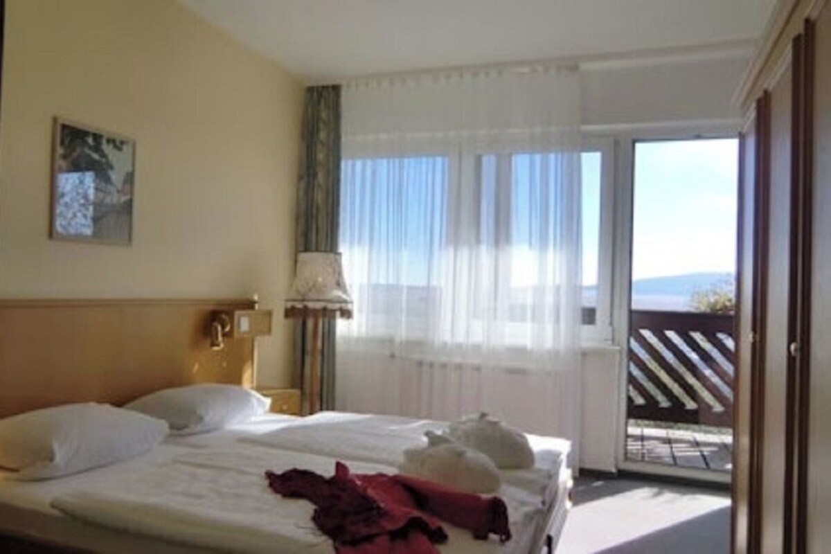 Ein Hotelzimmer mit Bett, Nachttisch, Lampe, einem Fenster mit Vorhängen und Blick ins Freie.