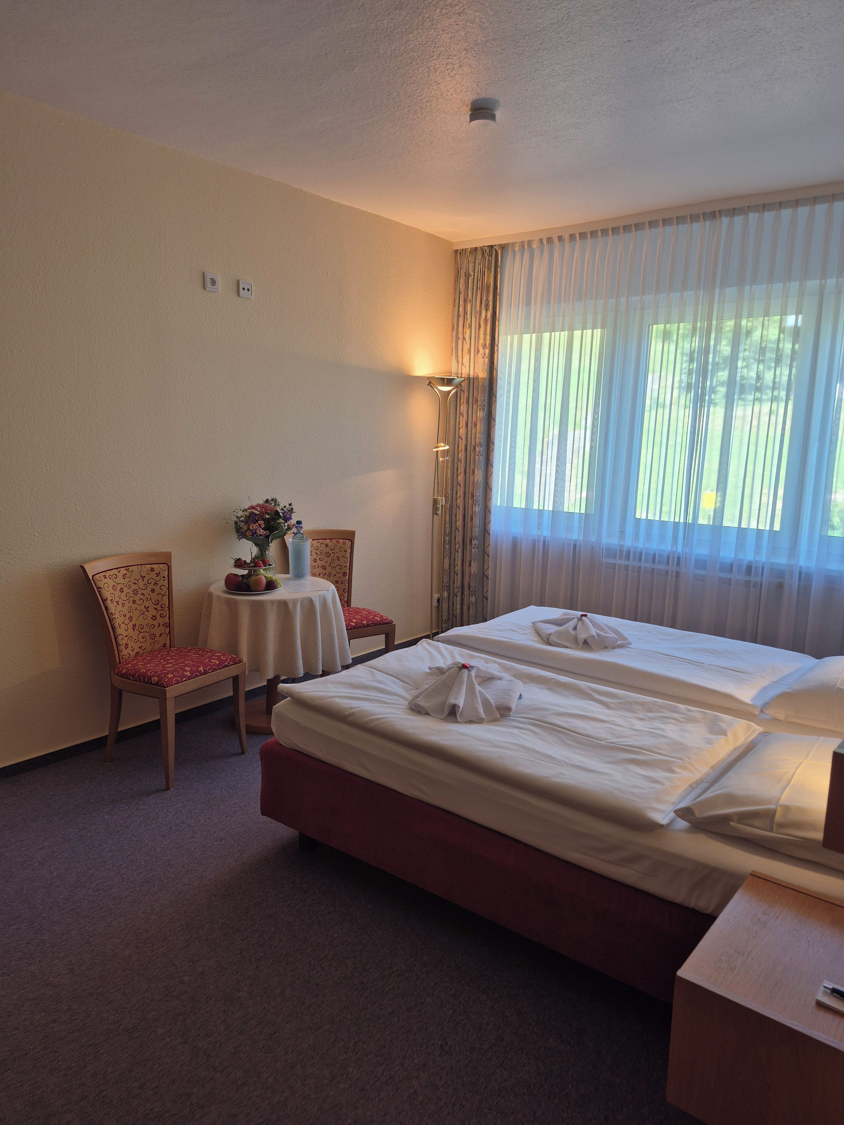 Habitación de hotel con dos camas, una silla, una mesita con un florero y una ventana con cortinas.