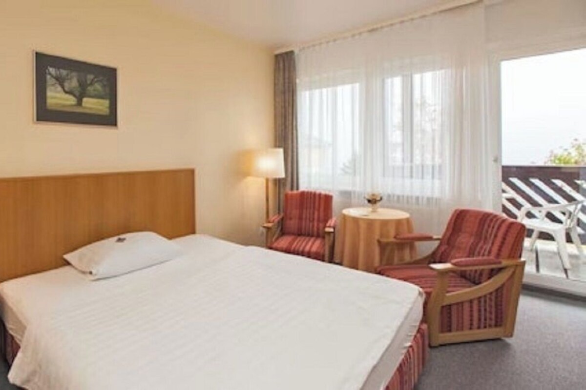 Ein Hotelzimmer mit einem großen Bett, einem Nachttisch mit Telefon, zwei Sesseln, einem kleinen runden Tisch und einem Fenster mit durchsichtigen Vorhängen.