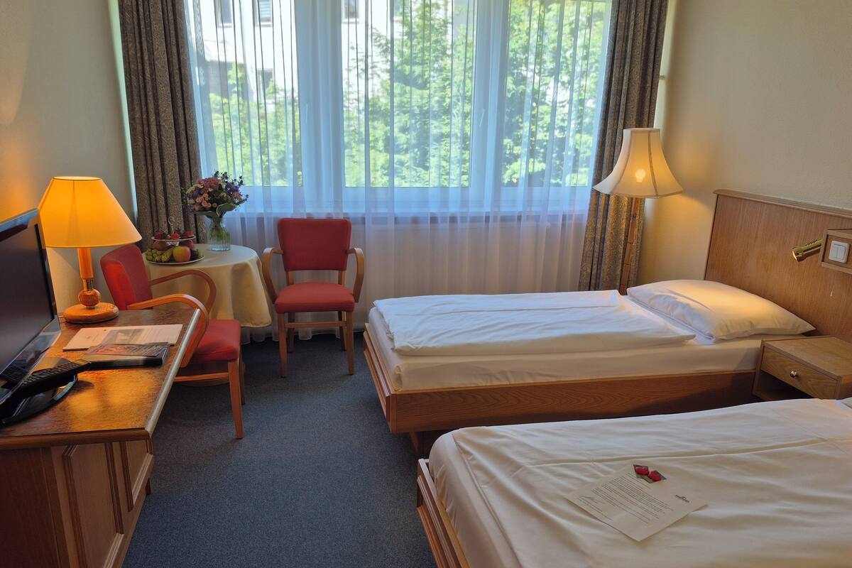Une chambre d’hôtel avec deux lits, un bureau avec une lampe, une chaise et une petite table sur laquelle est posé un vase de fleurs.