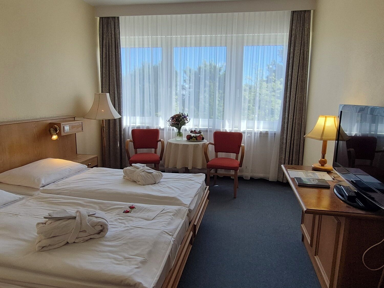 Ein Hotelzimmer mit zwei Betten, einem Schreibtisch mit Fernseher, einem kleinen Tisch mit Vase und Blumen sowie einem Fenster mit Vorhängen.