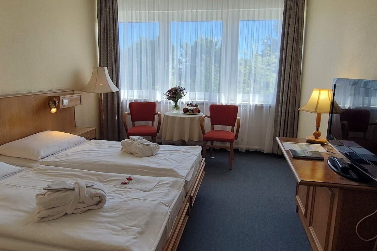 Une chambre d’hôtel avec deux lits, un bureau avec une télévision, une petite table avec un vase et des fleurs, et une fenêtre avec des rideaux.
