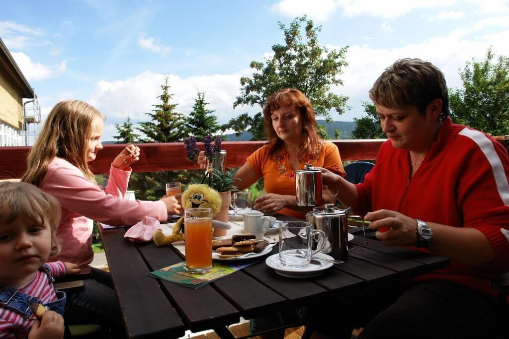 Eine Gruppe von Personen sitzt an einem Tisch im Freien mit einer Picknickausstattung, darunter Teller, Tassen und eine Kanne.