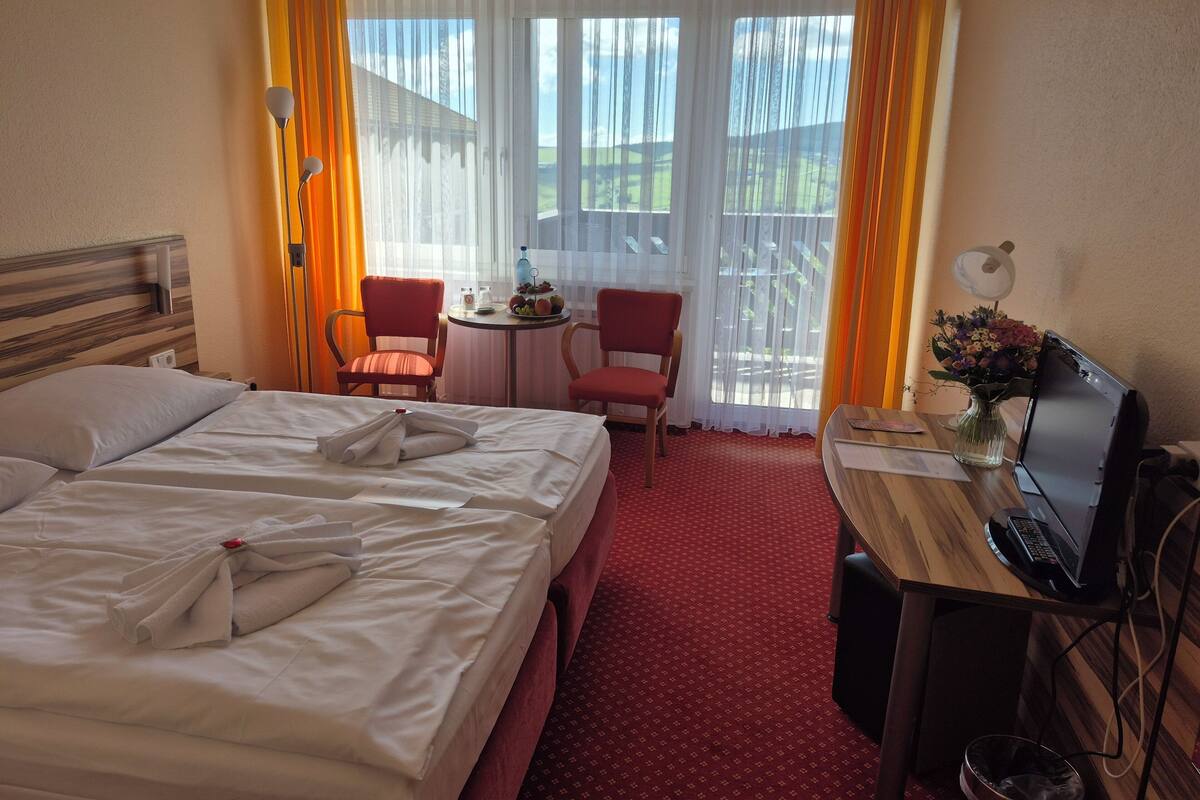 Une chambre d’hôtel avec un lit, deux chaises, une petite table sur laquelle se trouve un panier de fruits, un bureau avec un ordinateur portable et une fenêtre avec des rideaux.