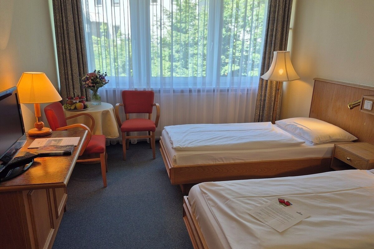 Habitación de hotel con dos camas, un escritorio con lámpara, una silla y una mesita con un jarrón de flores.