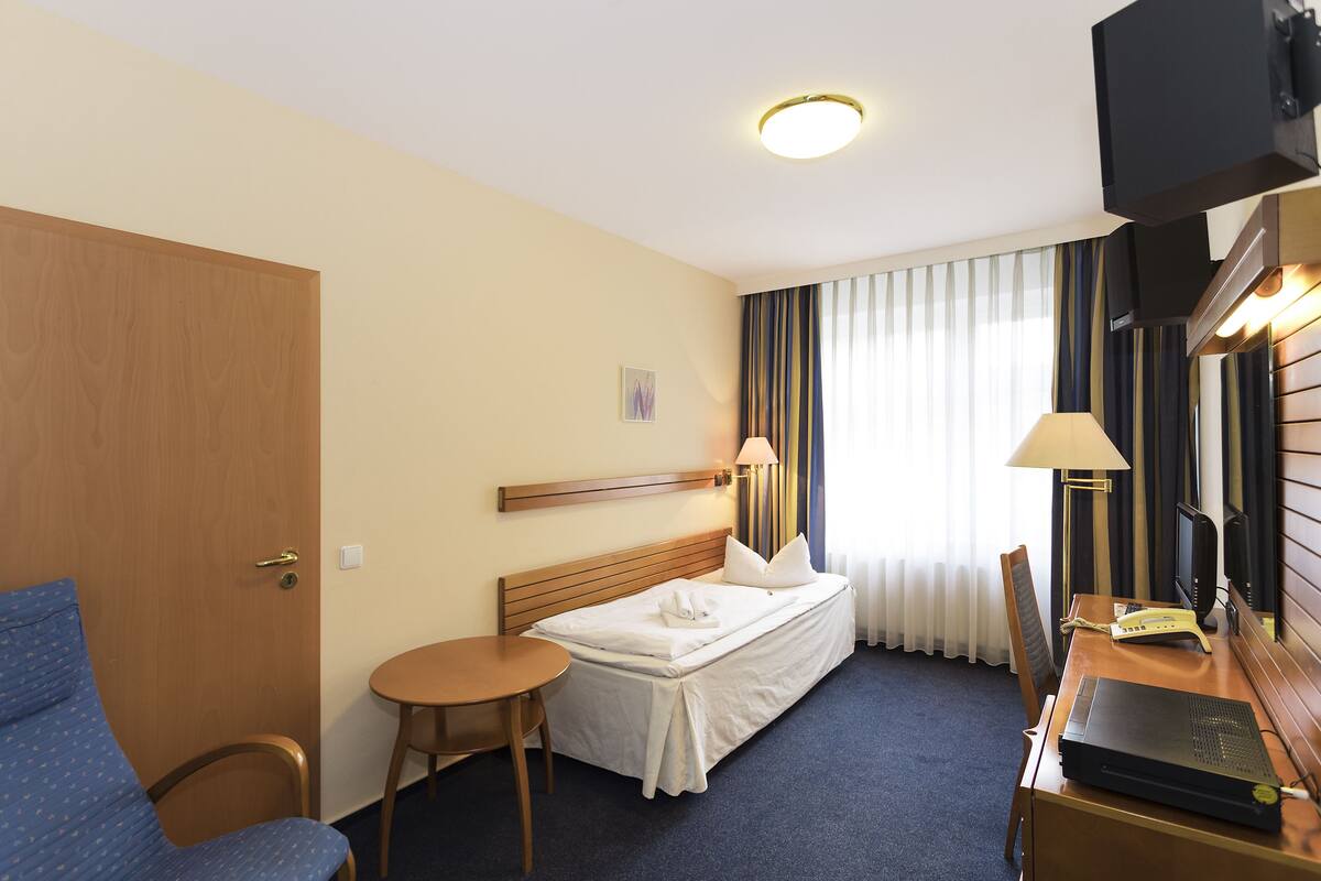 A hotel room with a bed, a desk with a laptop, a chair, a small table, and a TV.
