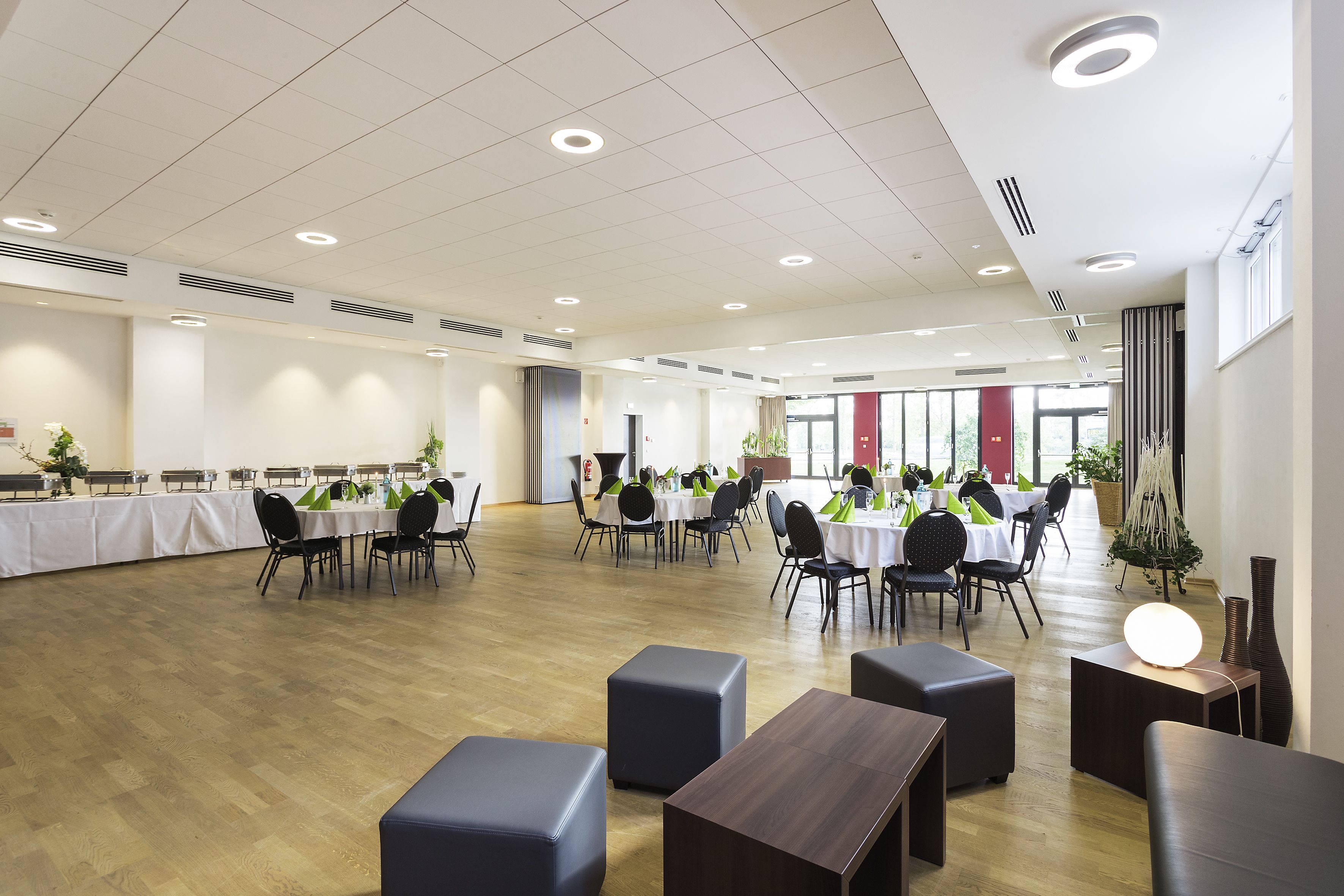 A spacious indoor dining area with multiple round tables and chairs, a wooden floor, and ceiling lights.