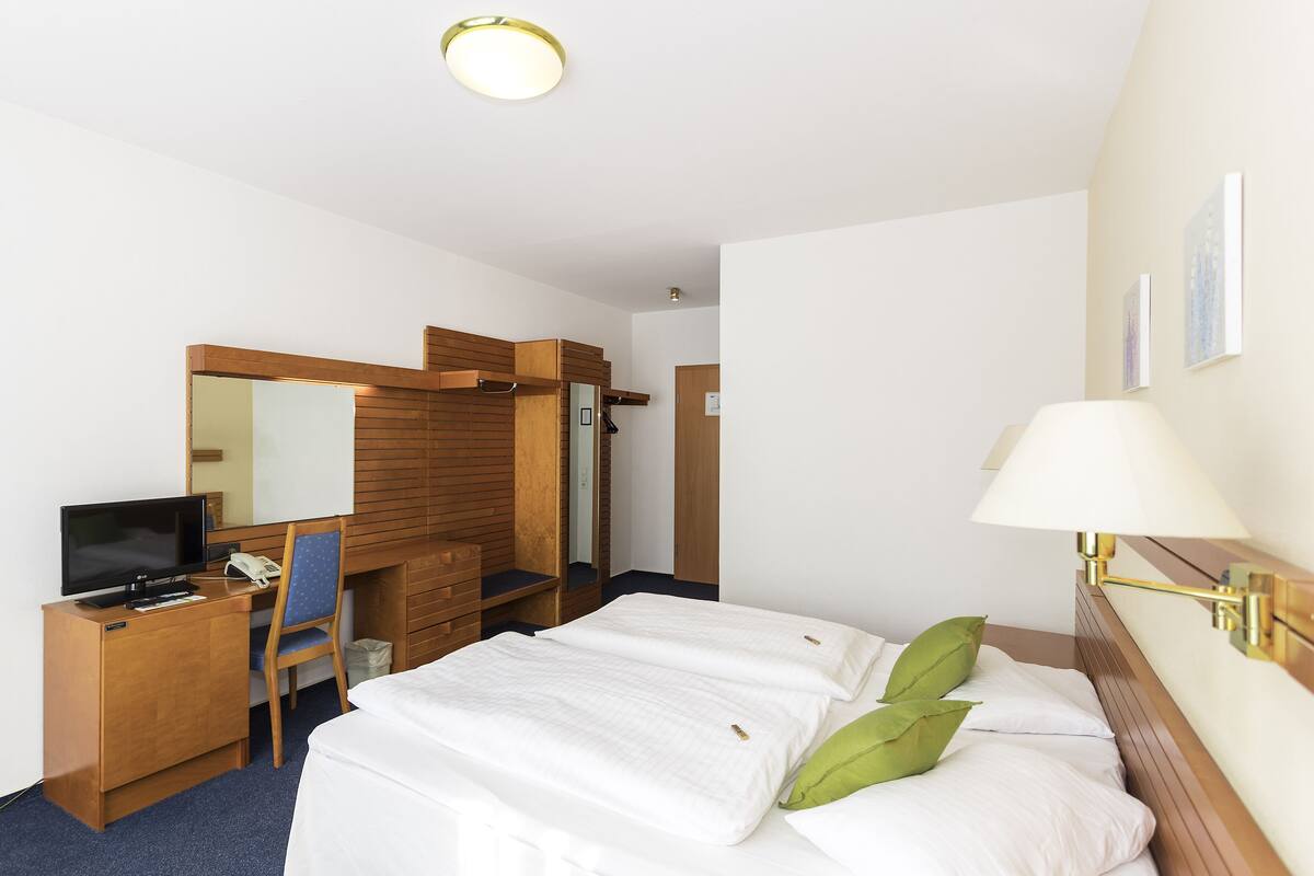 A hotel room with a bed, a desk with a computer, a chair, a wardrobe, and a lamp.