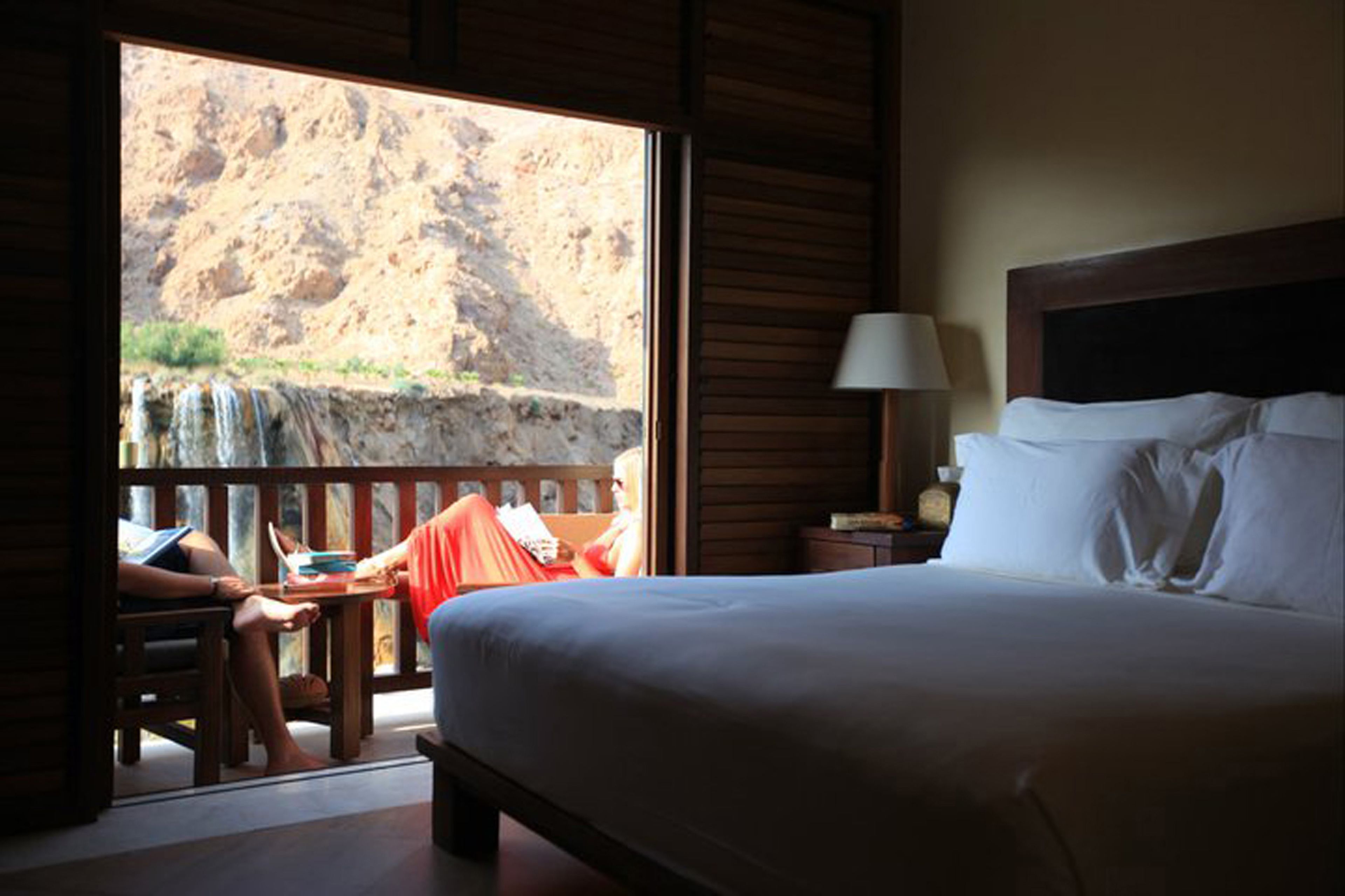 A bedroom with a bed, a nightstand, a lamp, and a view of a rocky landscape through a window.