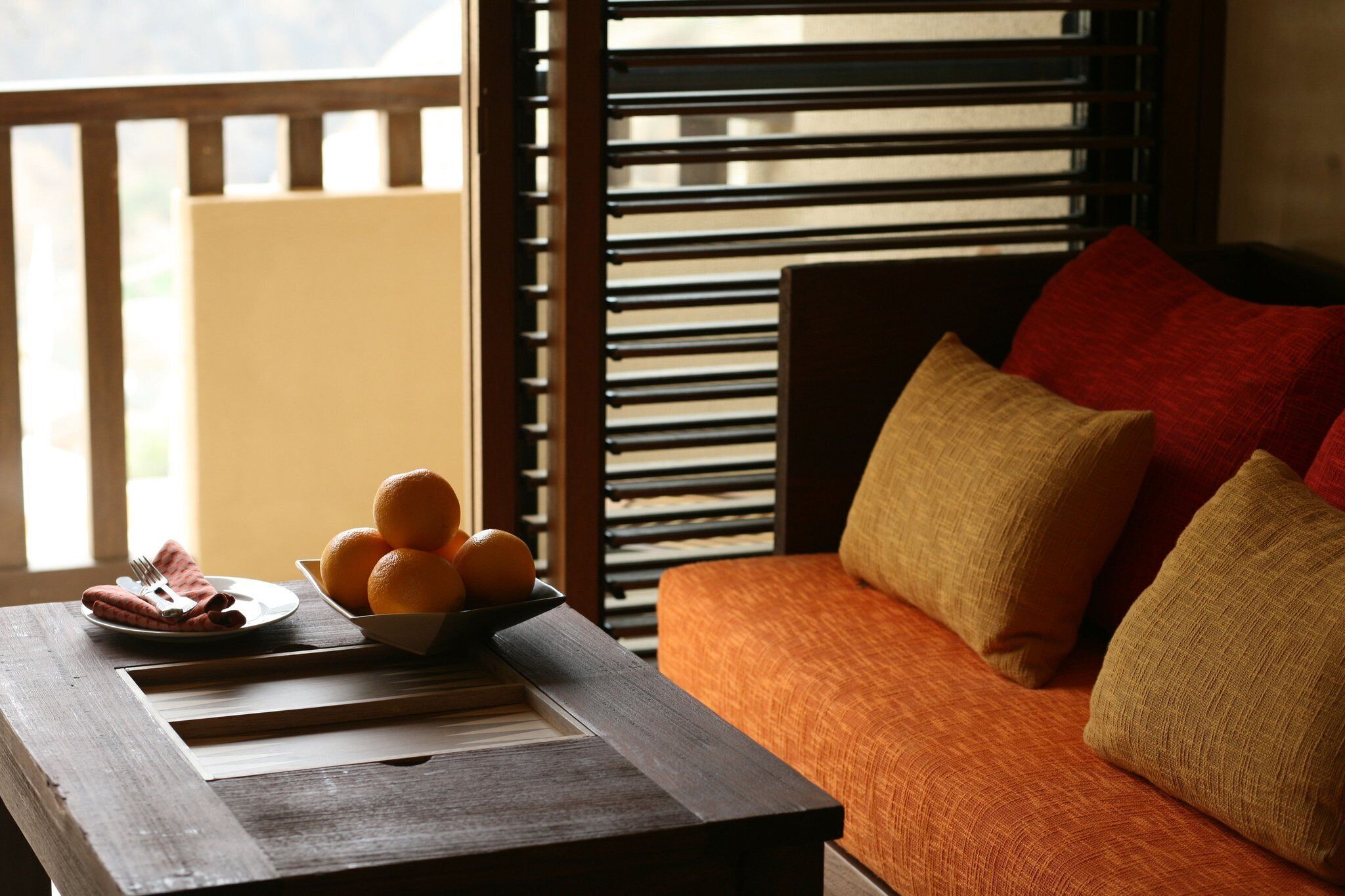 Una mesa de madera con un plato de naranjas y un bol de fruta, un sofá con cojines naranja y una ventana con persianas horizontales.