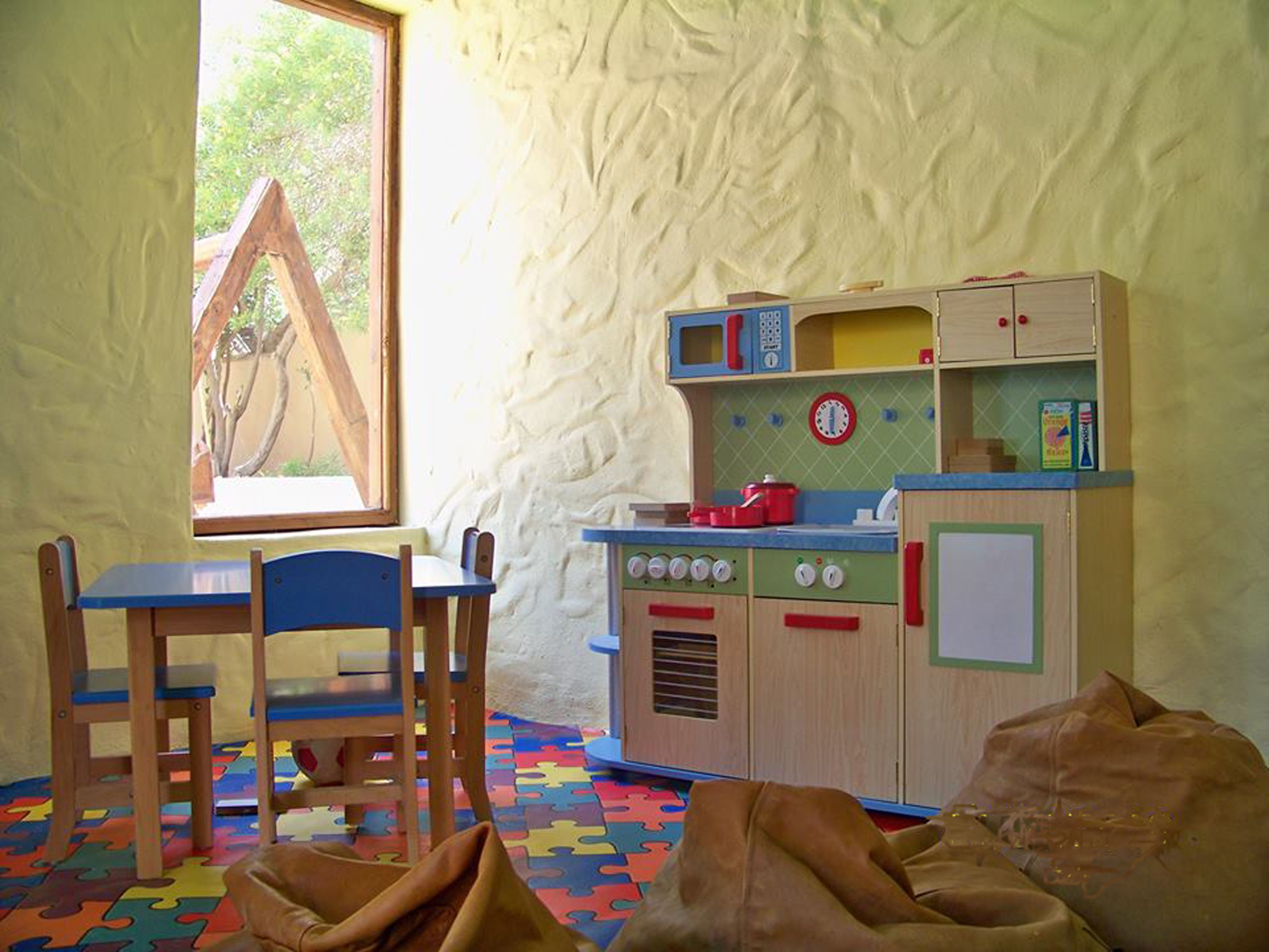 Una sala giochi con un tavolo e sedie in legno, una cucina giocattolo e una finestra vista alberi.