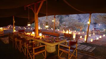 An outdoor dining area with a long table set for multiple people, surrounded by chairs, and illuminated by numerous candles and lanterns.