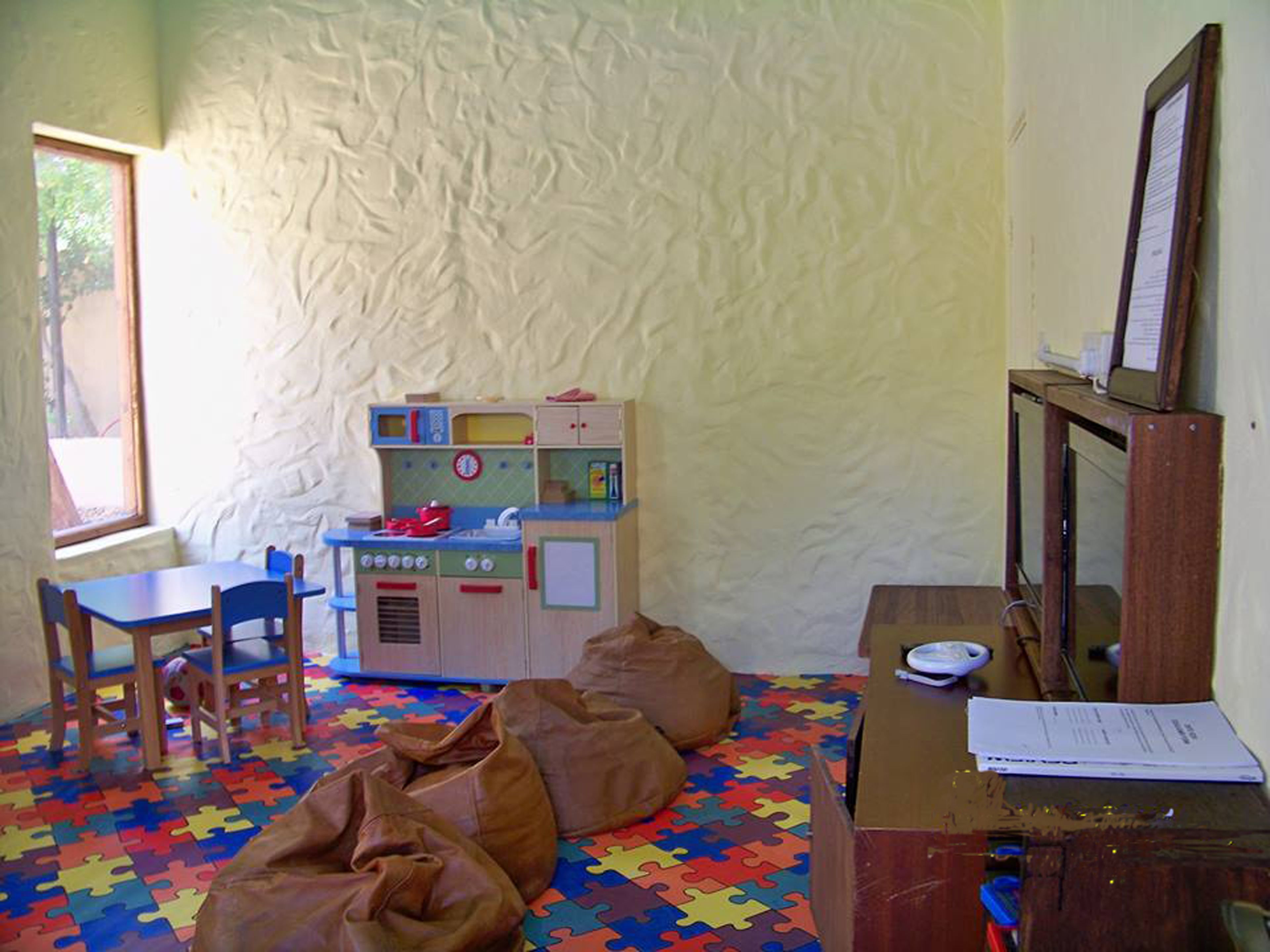 A room with a play kitchen, a wooden desk, a chair, bean bags, and a puzzle floor.