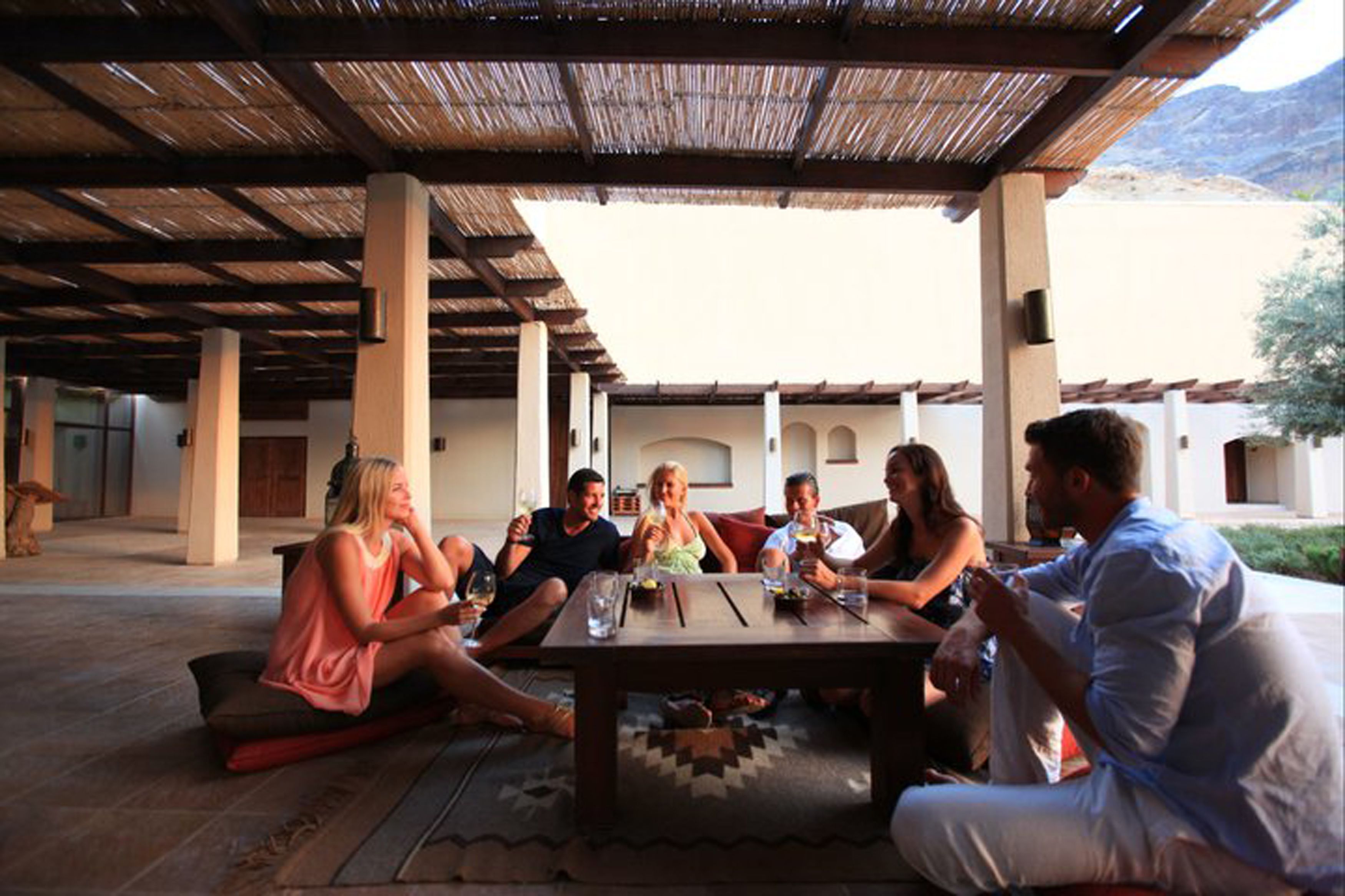 A group of people seated around a wooden table in an outdoor covered area with a thatched roof, surrounded by pillars and a mountainous backdrop.