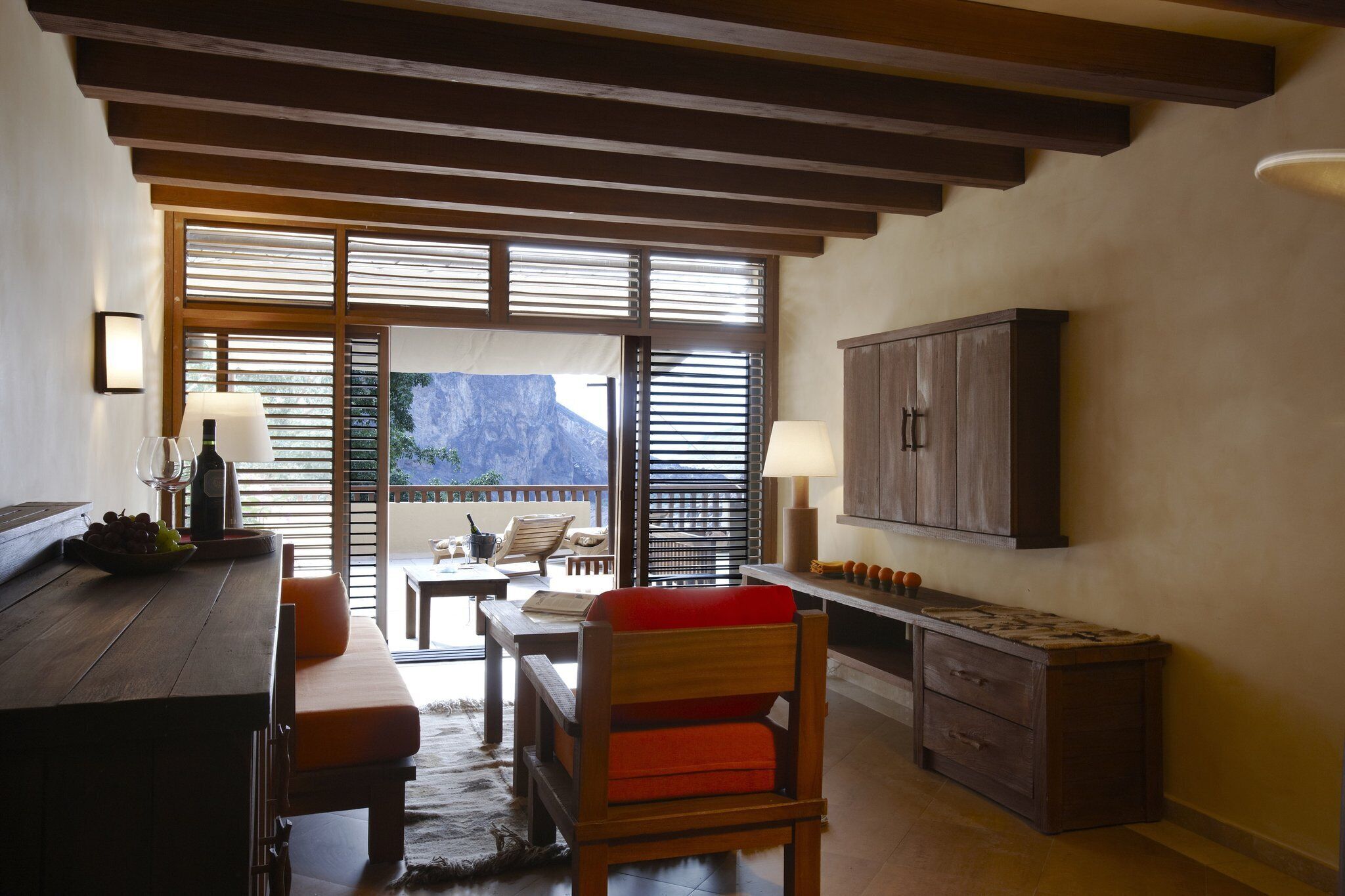 A cozy room with a wooden bench, a table, and a cabinet. There is a window with blinds, and a view of a mountain is visible outside.