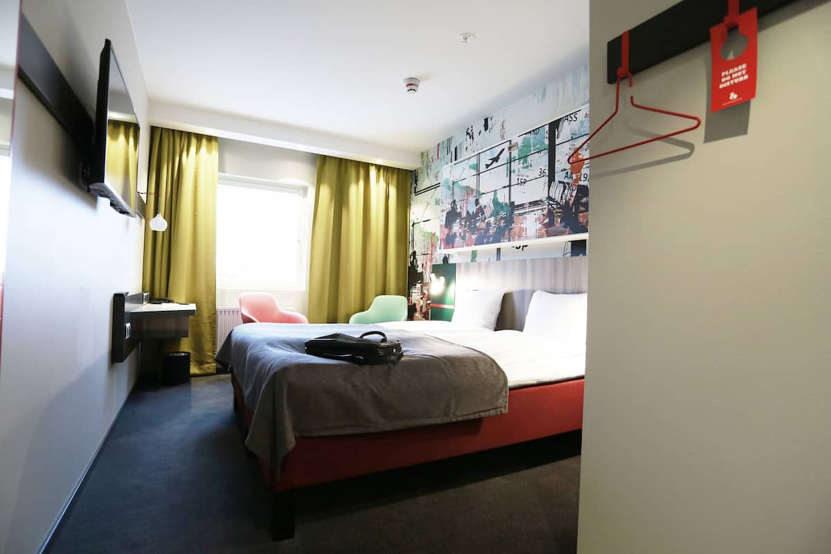 A hotel room with a bed, a desk, a chair, a TV, and a wall with artwork.