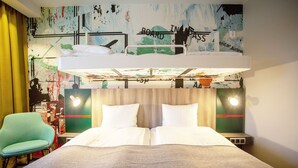 Ett hotellrum med en våningssäng, en grön stol och en vägg med abstrakt konst.