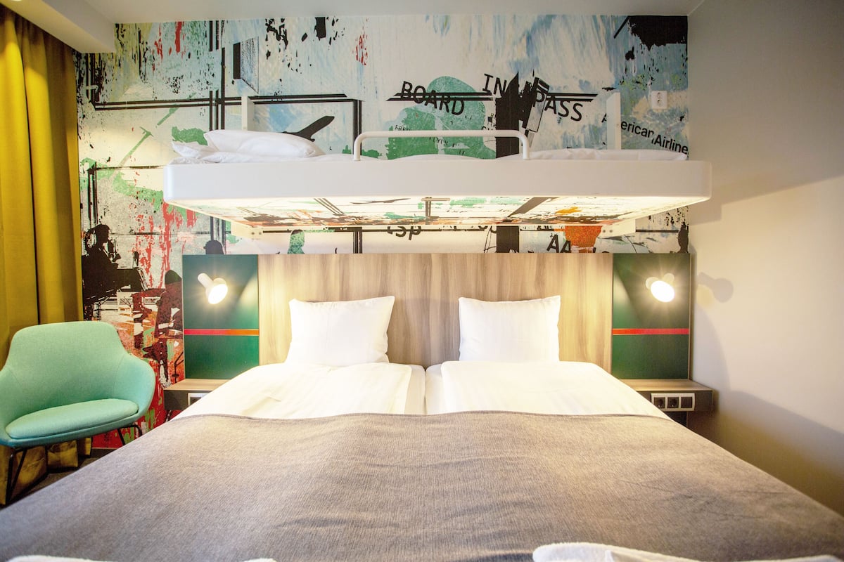 A hotel room with a bunk bed, a green chair, and a wall with abstract art.
