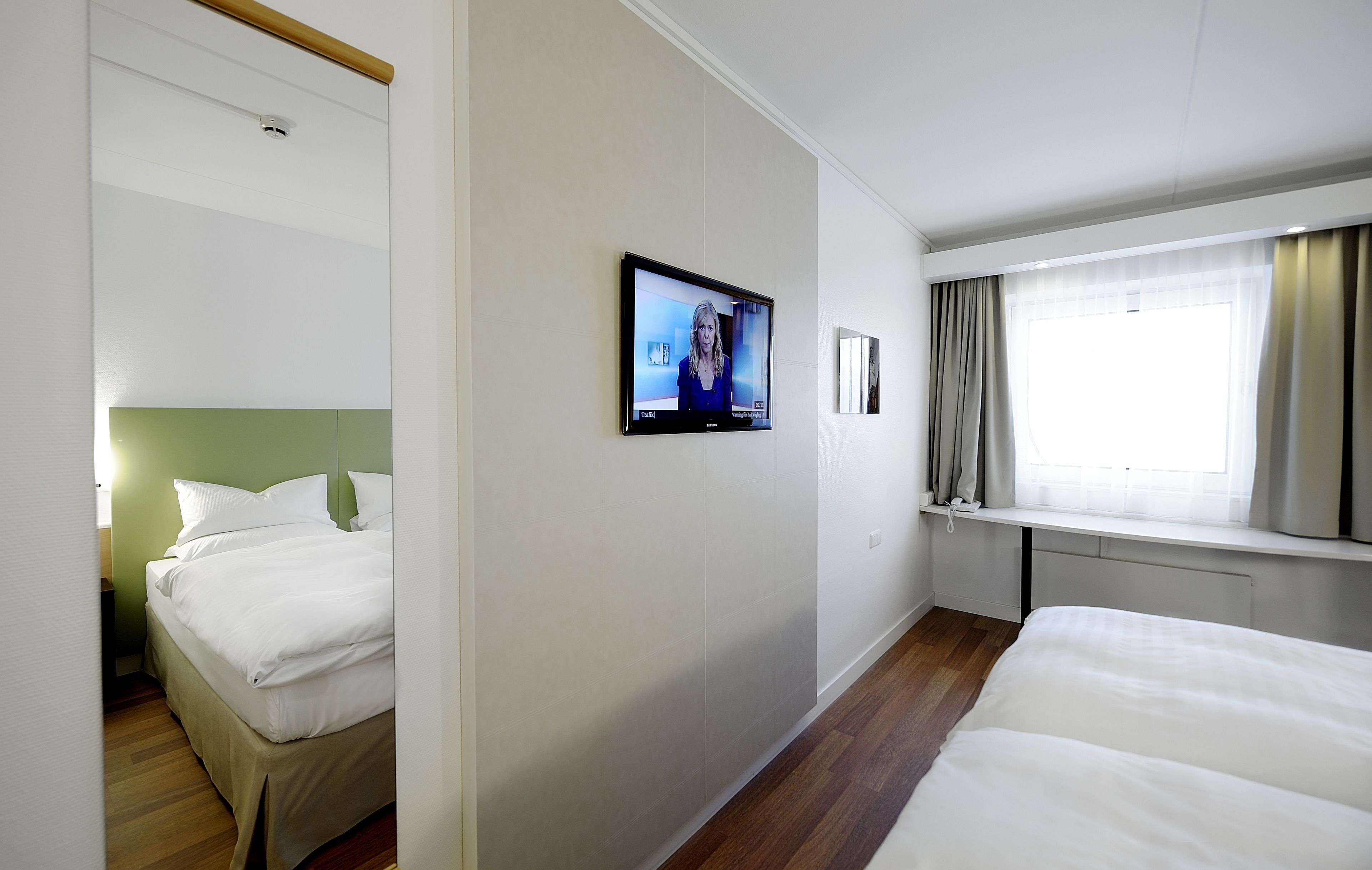 Ett hotellrum med en säng, en tv som är monterad på väggen, ett fönster med gardiner och ett nattduksbord.