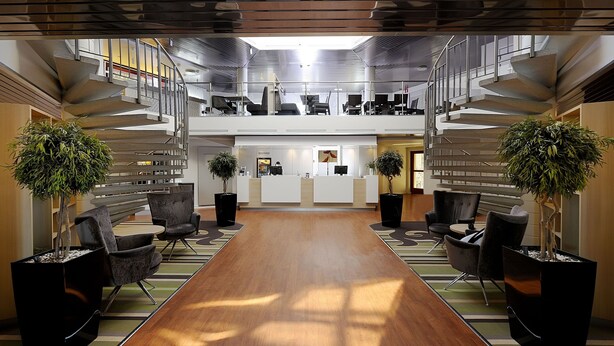En rymlig entréhall med en central receptionsdisk, två sittgrupper och en modern trappa.
