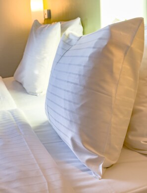 Una cama bien hecha con sábanas y almohadas blancas, una lámpara de noche y una pared verde de fondo.
